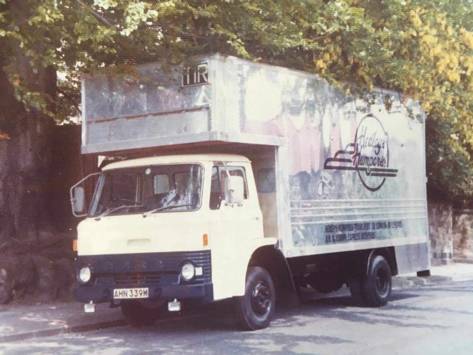 A vintage delivery truck with a white cab and black bumper, parked on a street surrounded by trees.