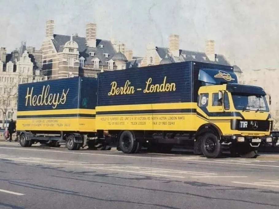 A yellow and blue delivery truck with 'Hedley's' and 'Berlin - London' written on the side, parked on a city street with historic buildings in the background.
