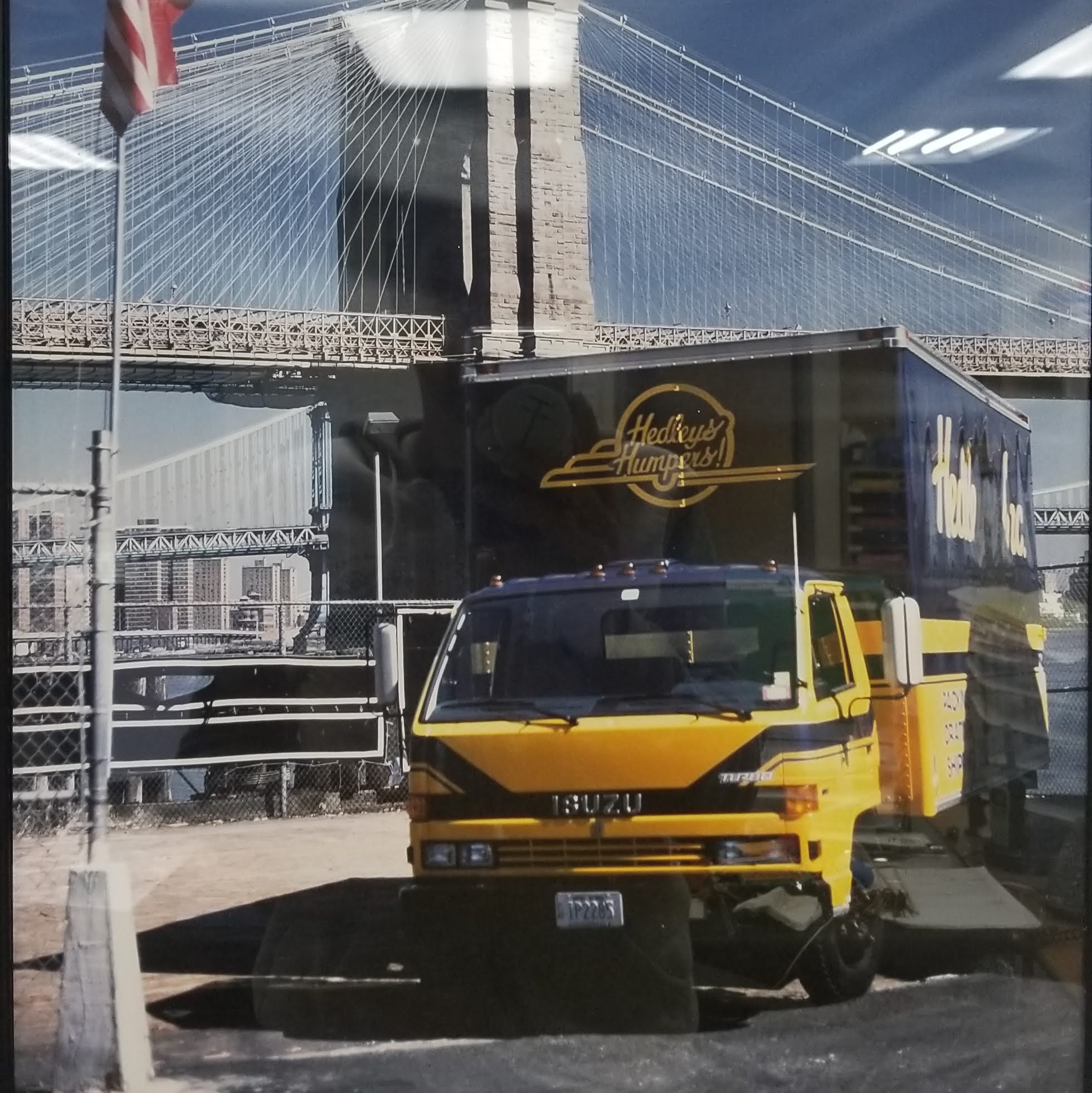 A yellow and black Isuzu service truck with a box on the back, parked behind a chain-link fence with the Brooklyn Bridge in the background, and the logo 'Hedley's Hampers!' on the box.