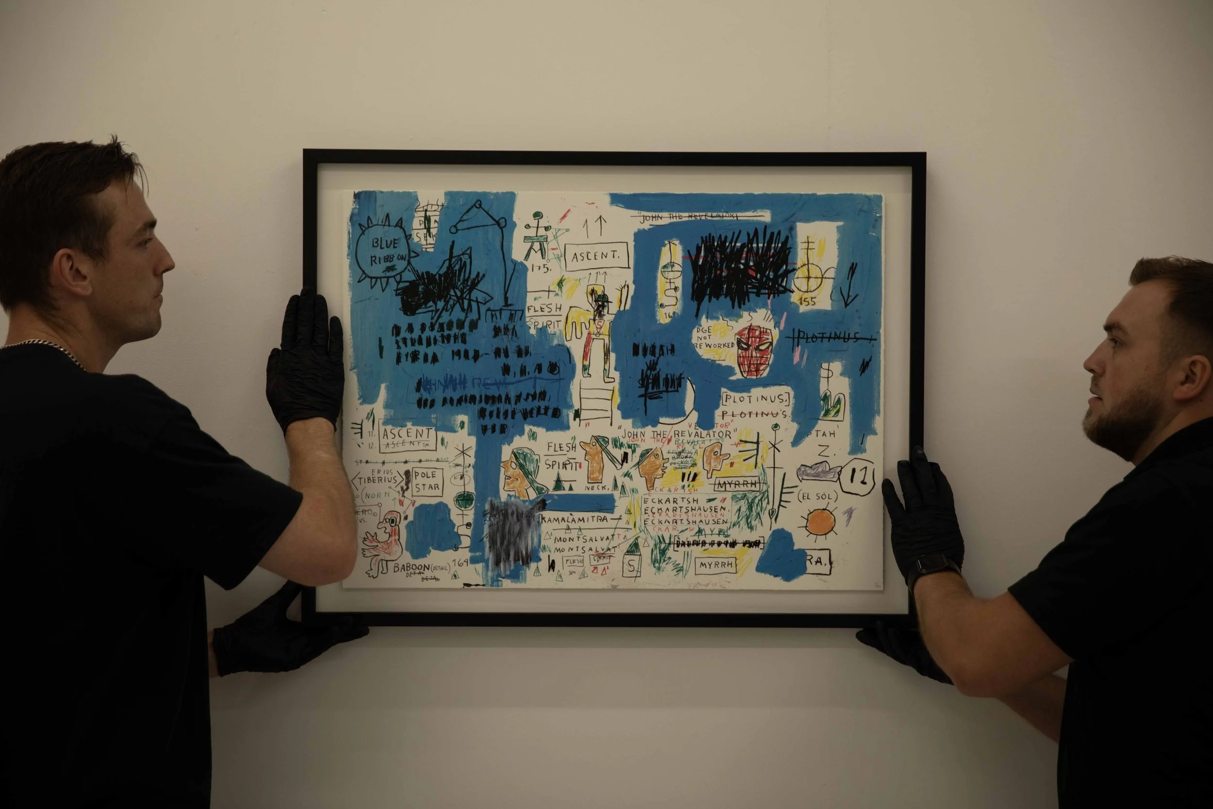 Two men with black gloves handling a framed abstract artwork on a white wall. The artwork features sketches, text, and colorful illustrations in blue, red, yellow, green, and black.