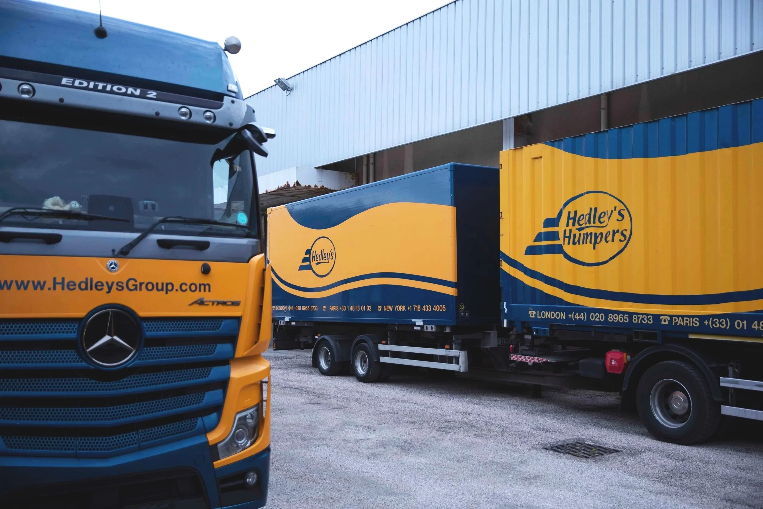 Two large trucks with Hedley's Humpers branding parked outside a warehouse. One truck is partially visible in the foreground, while the other is in the background. The trucks are blue and yellow.