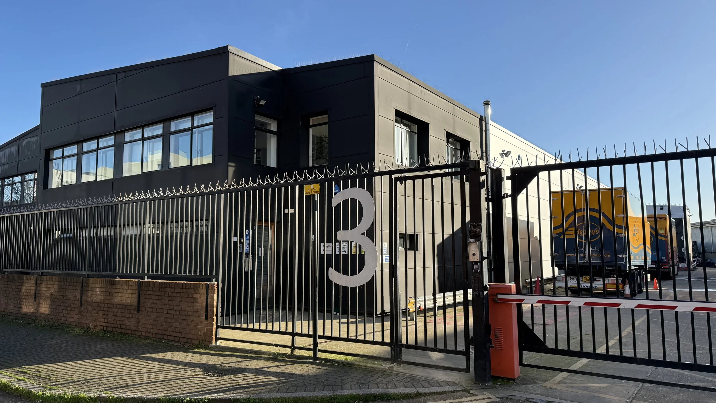 A modern two-story commercial building with a black upper section and large windows, enclosed by a tall black metal fence with a gate marked with the number 3, and trucks parked behind the fence in a parking lot.