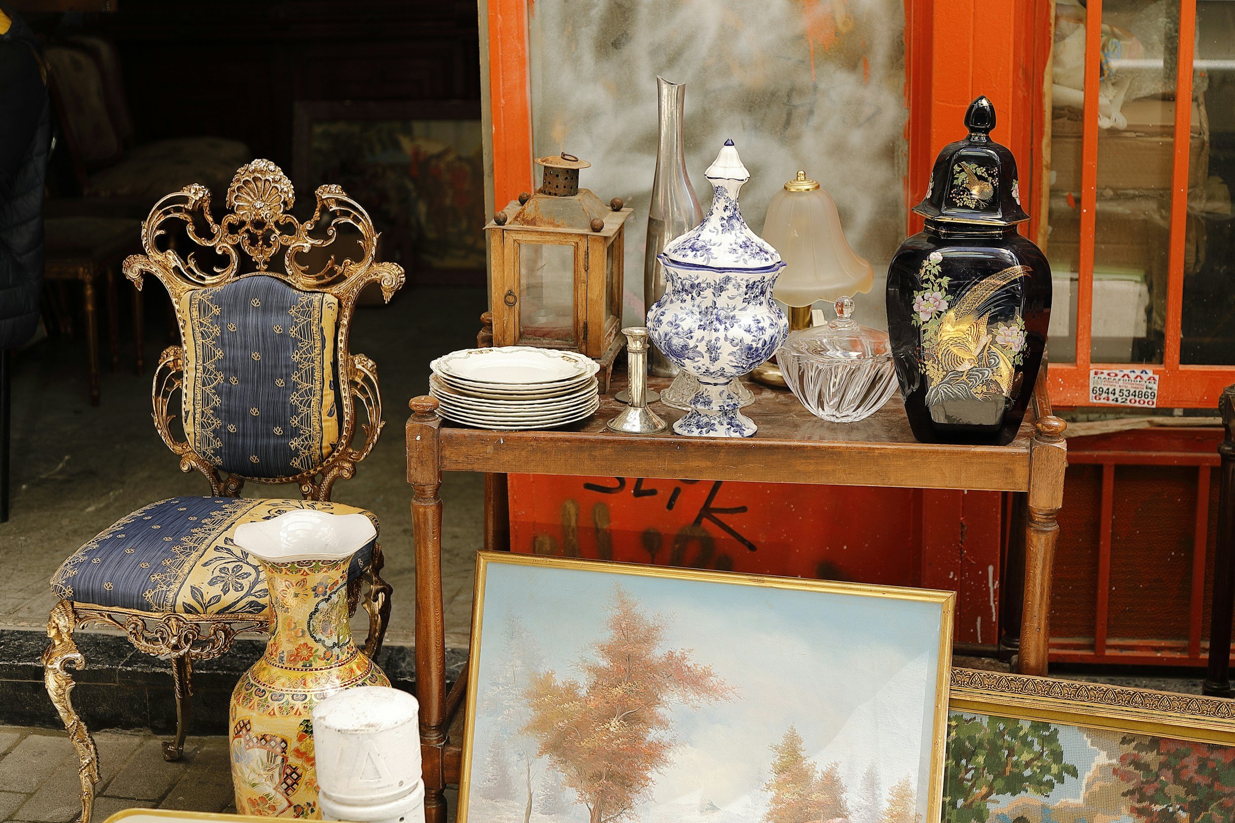 Une table en bois avec des verres, lampes en céramique et autres objets décoratifs. Un fauteuil orné de dorures et de tissu bleu et or est à côté, avec une grande vase en céramique à motifs. Deux tableaux de paysage sont posés au sol, devant la table.