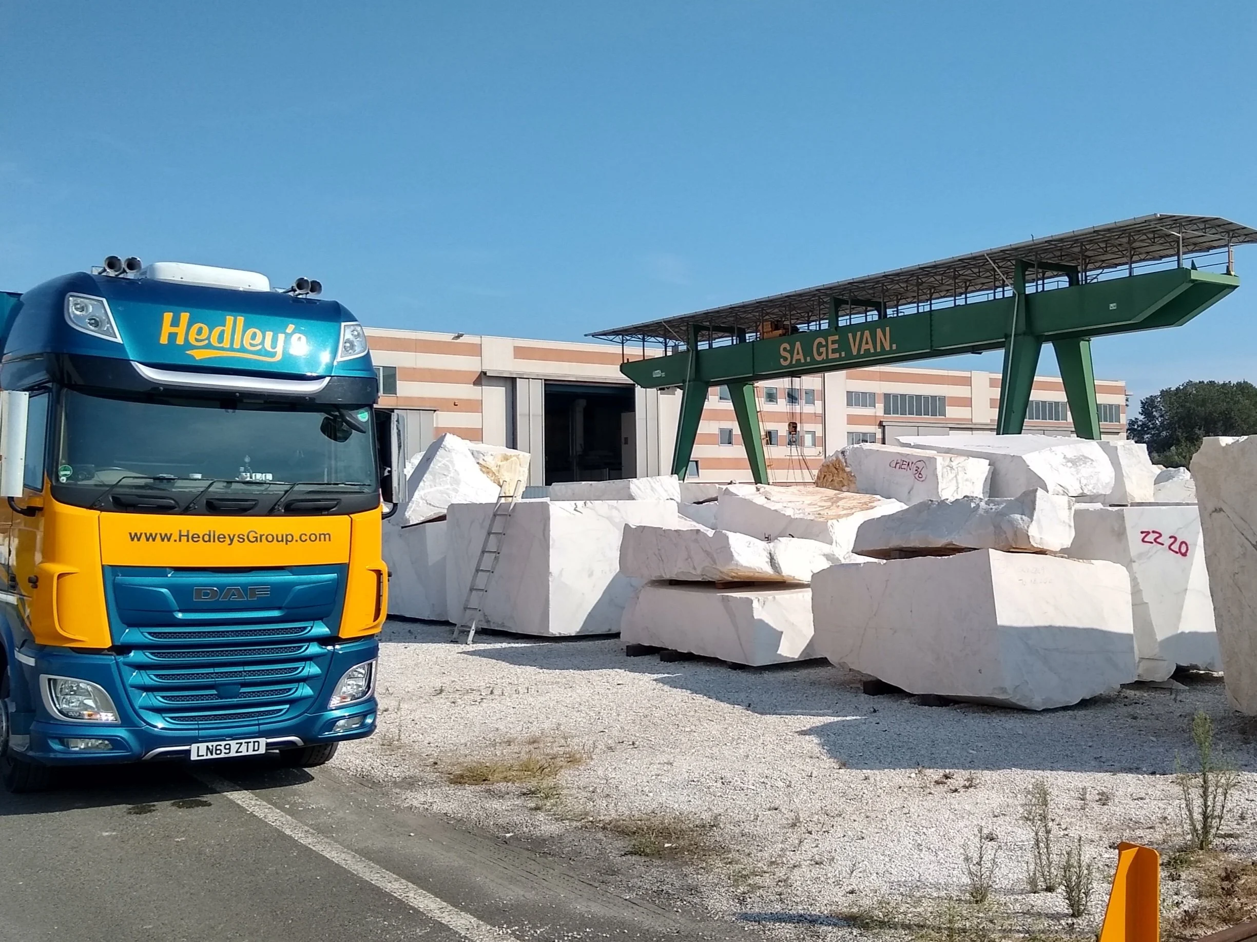 Un camion Mercedes-Benz jaune et bleu bloquant une petite route étroite dans un village européen, entouré de maisons en bois et de végétation.