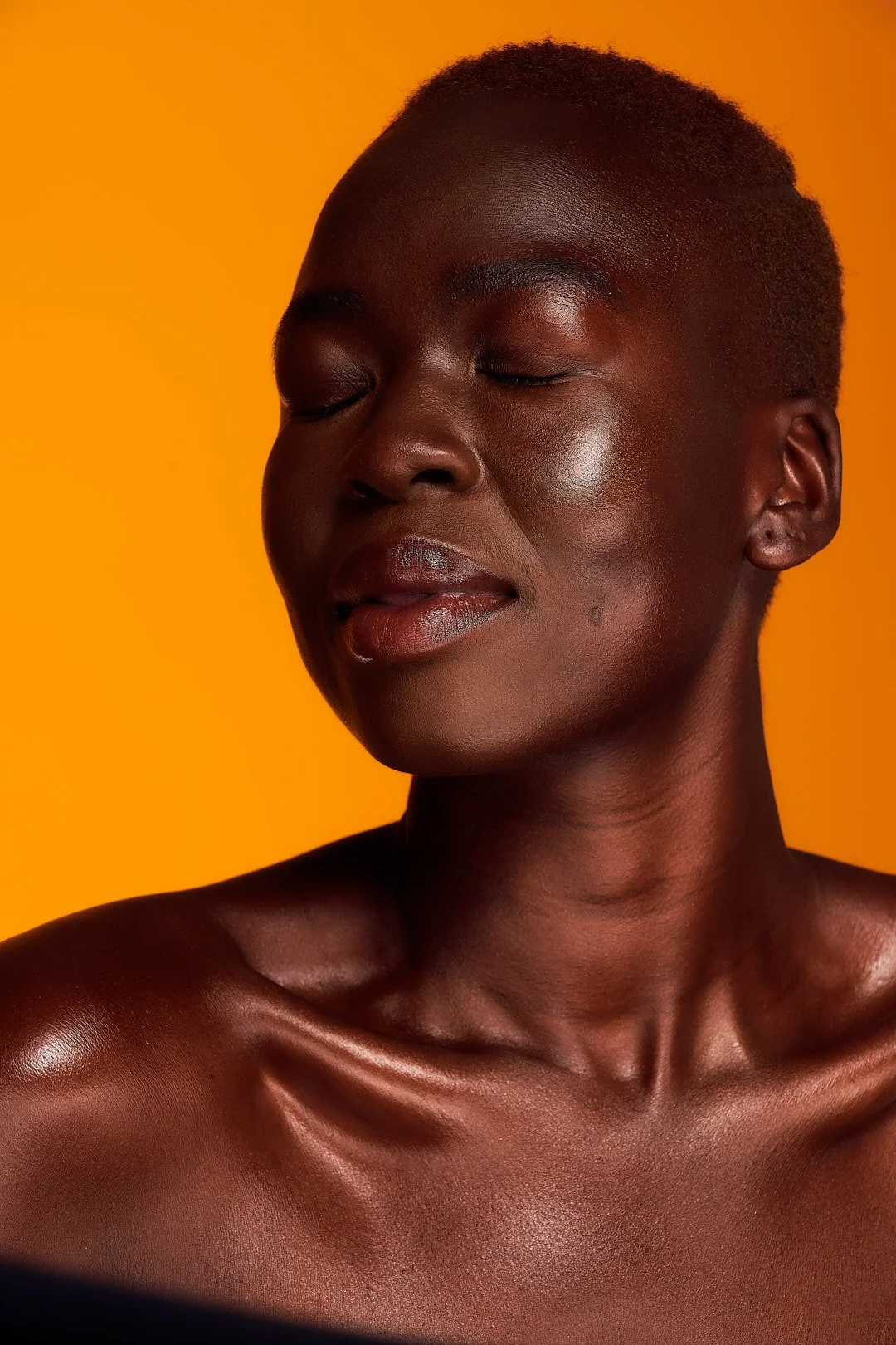 A close-up portrait of a woman with dark skin and short hair, eyes closed, on a bright orange background.