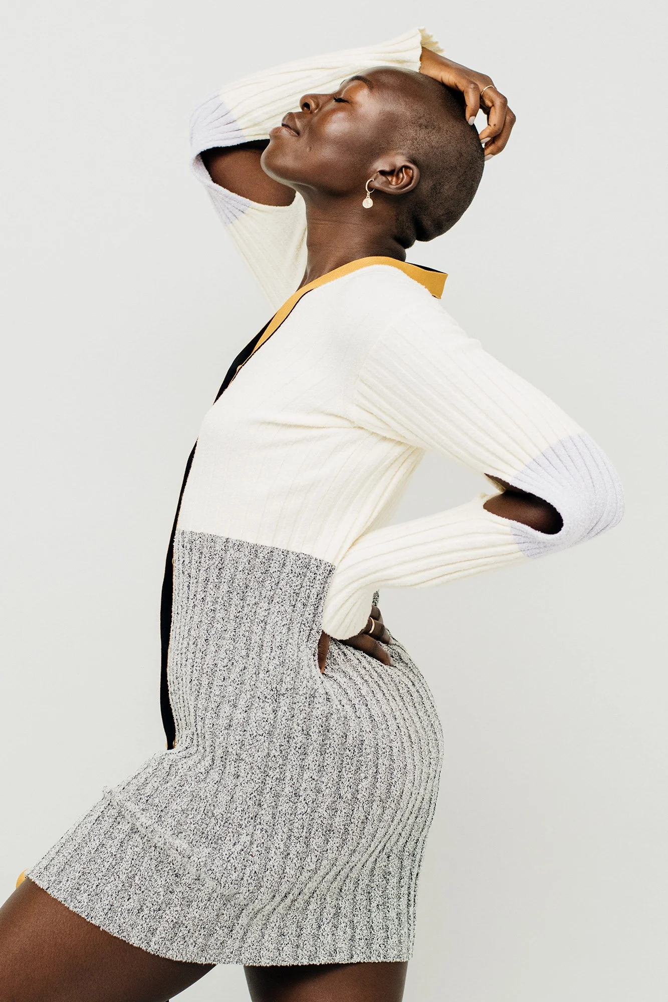 A woman with dark skin and a buzz cut hairstyle poses with her hand resting on her head and her eyes closed. She is wearing a vertically striped, two-tone sweater and matching skirt, against a plain light background.