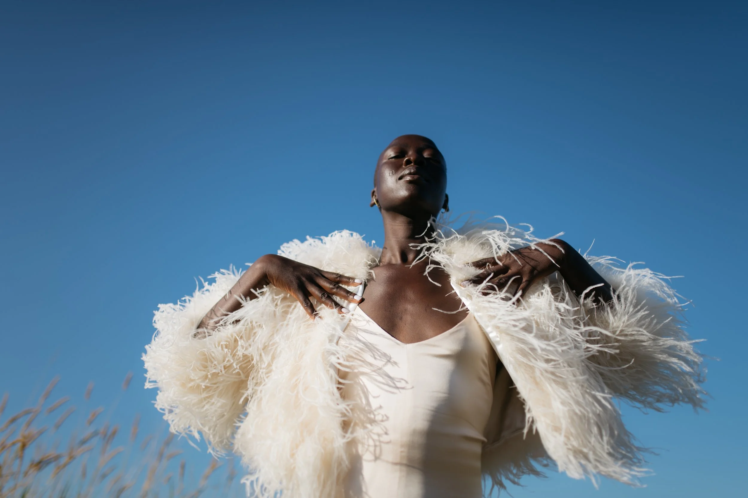 A woman with dark skin in a white dress and feathered cream-colored coat standing outdoors under a clear blue sky, looking confidently at the camera with hands on her shoulders.
