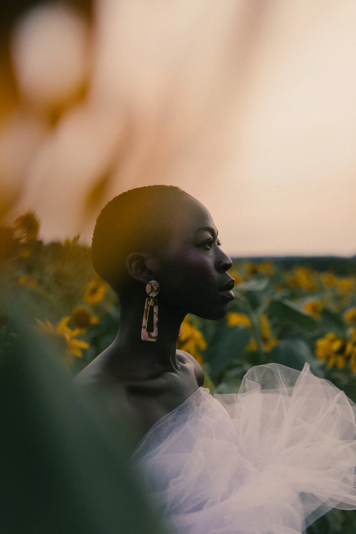 A woman with dark skin and short hair, wearing large earrings and a sheer, white, tulle dress, standing among sunflowers during sunset.
