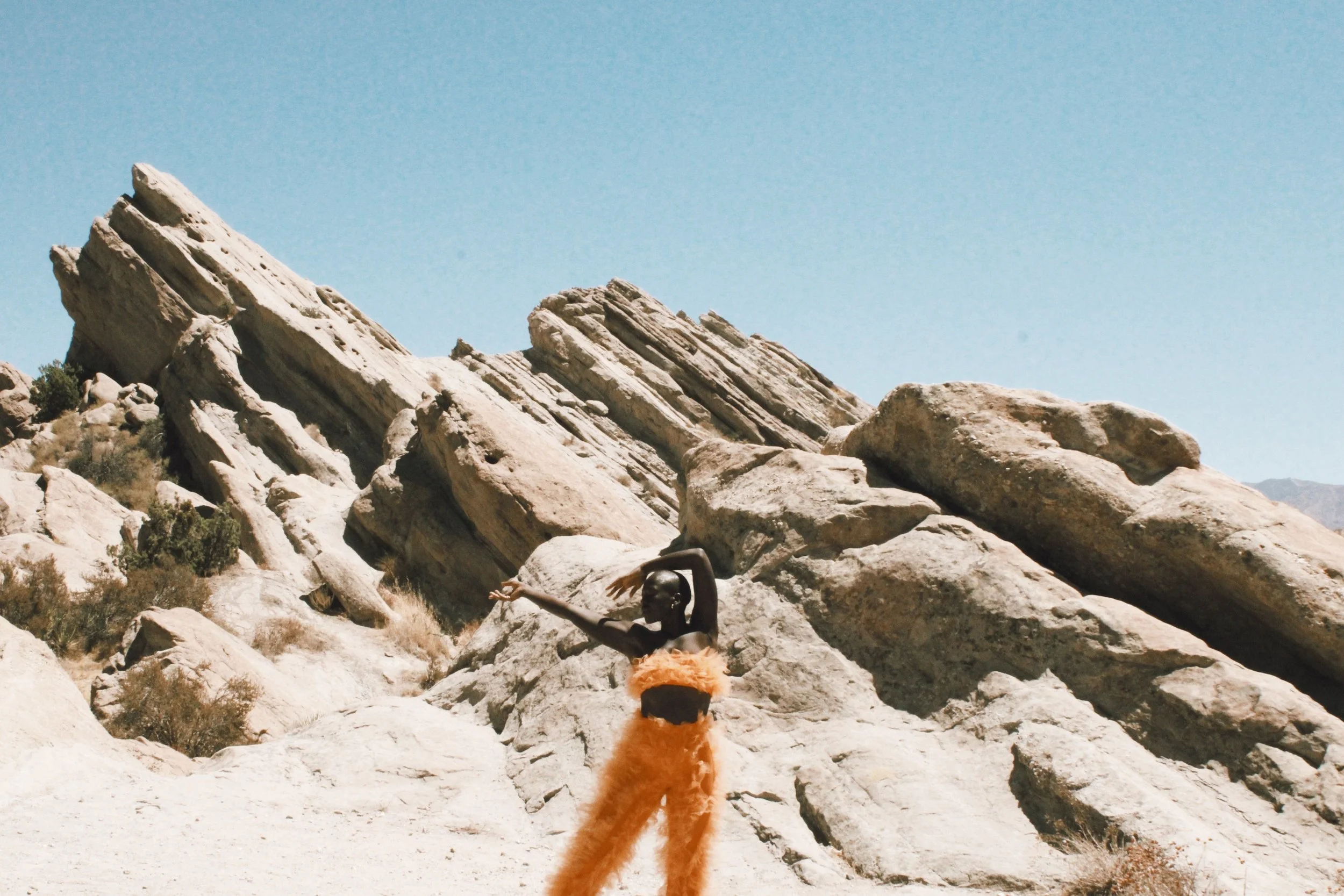 A person with dark skin wearing orange furry pants and a black top, standing in a desert landscape with large rock formations and a clear blue sky, striking a dance pose.