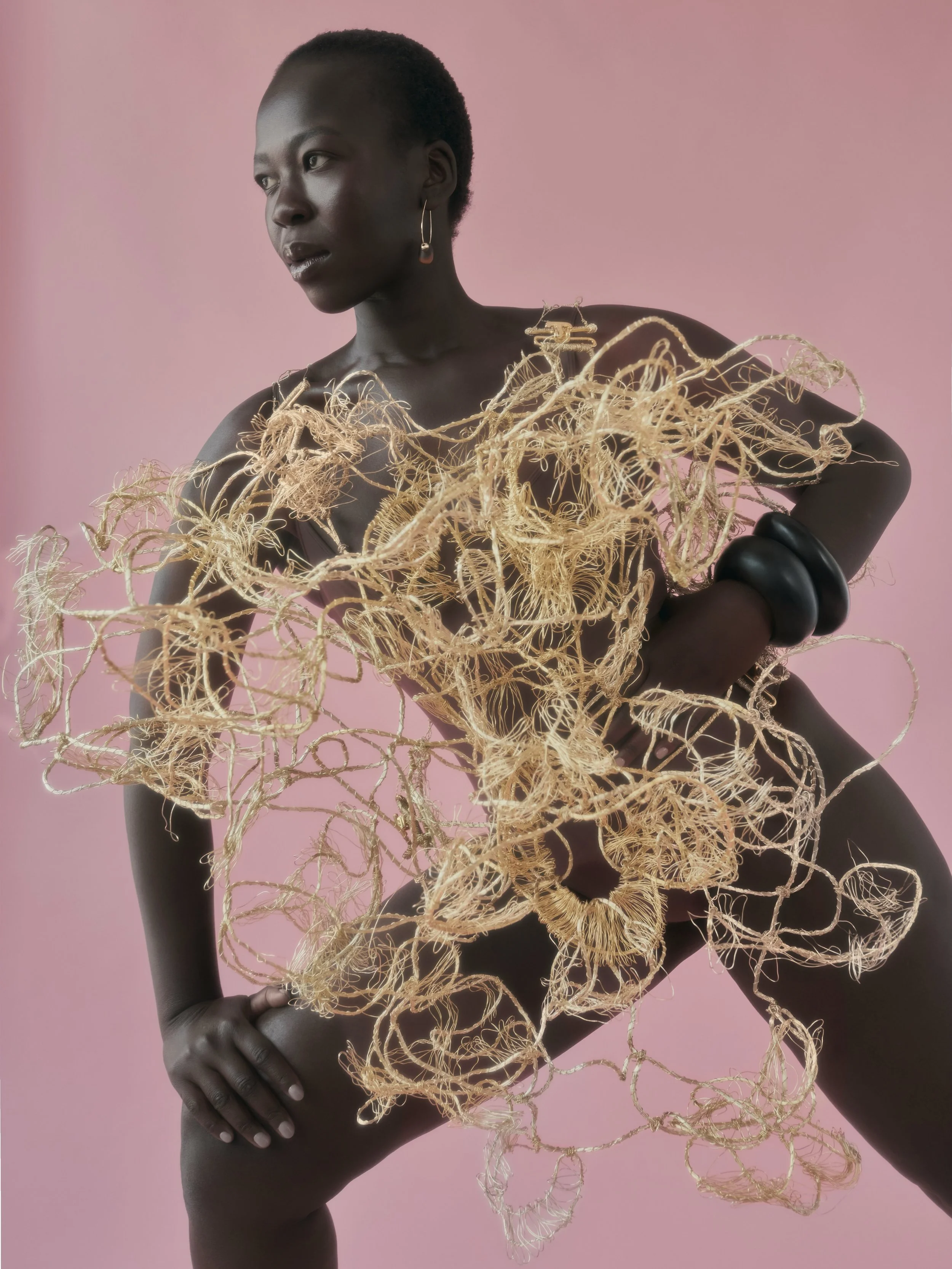 Woman with short hair and dark skin posed against a pink background, holding intertwined golden wire sculpture covering her torso and hips, wearing black bangles and earrings.