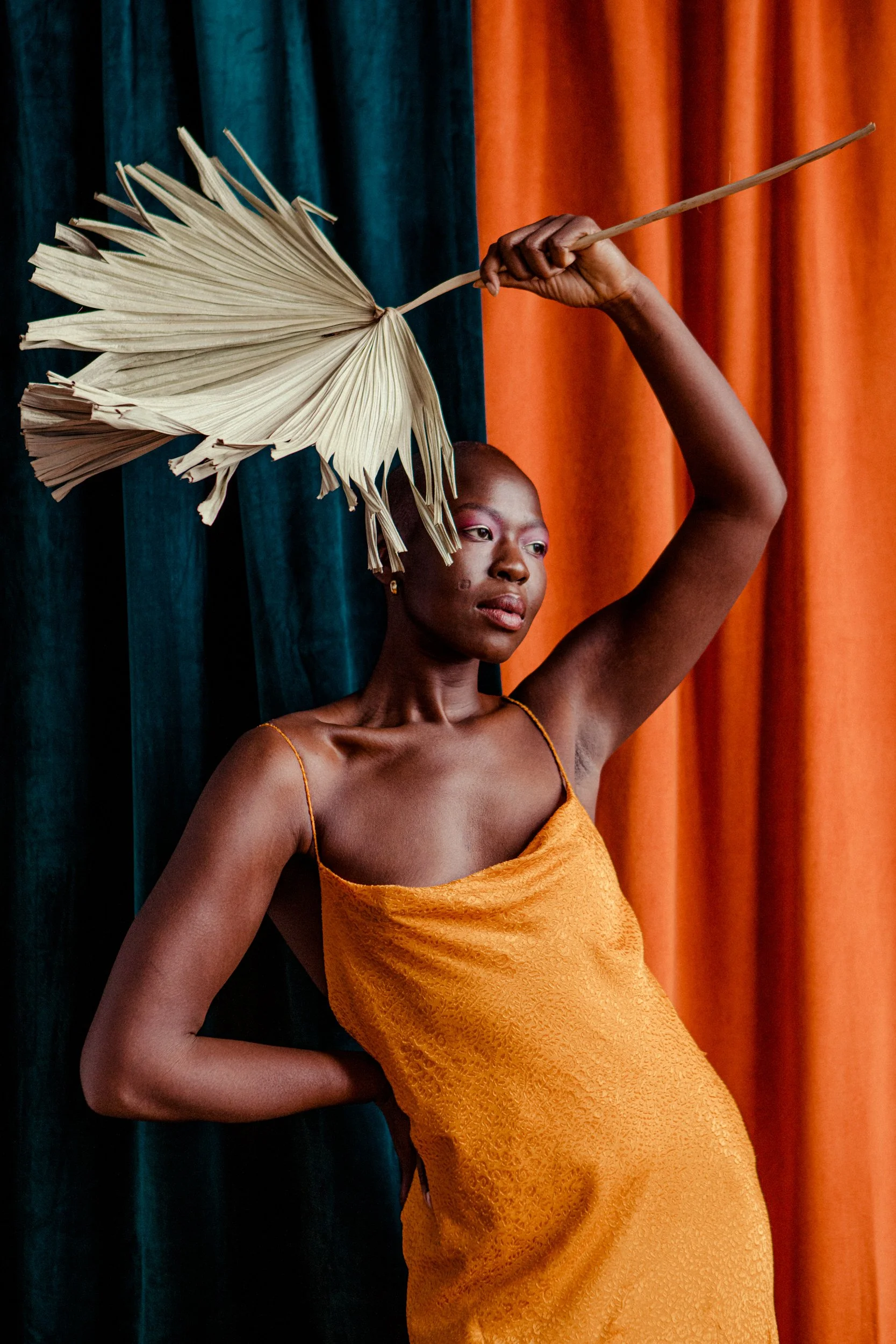 A woman with dark skin in a yellow dress holding a dried palm leaf above her head, standing against colorful curtains.