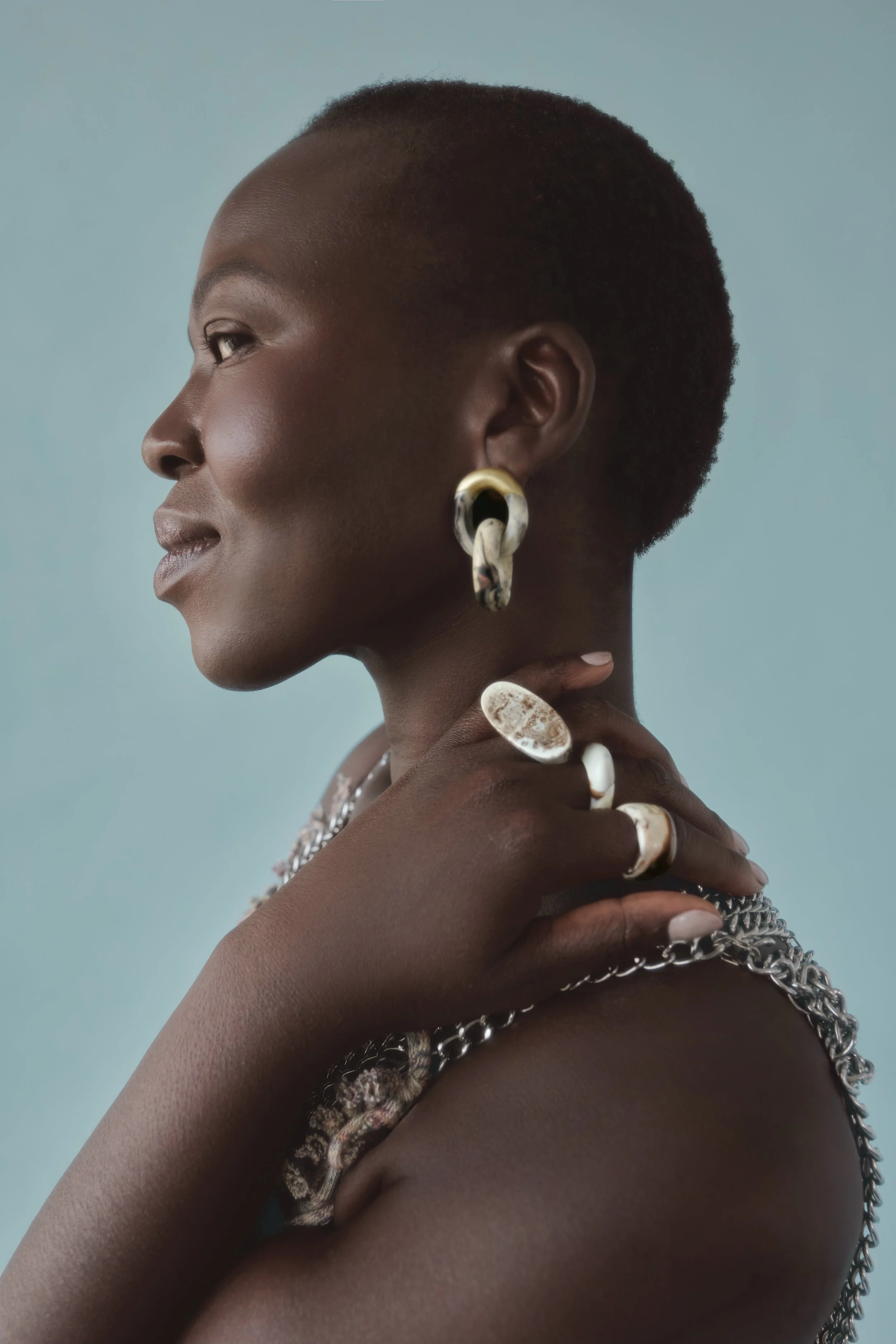 Side profile of an African American woman with short hair, wearing silver and gold jewelry including earrings, rings, a necklace, and a chain, against a light blue background.