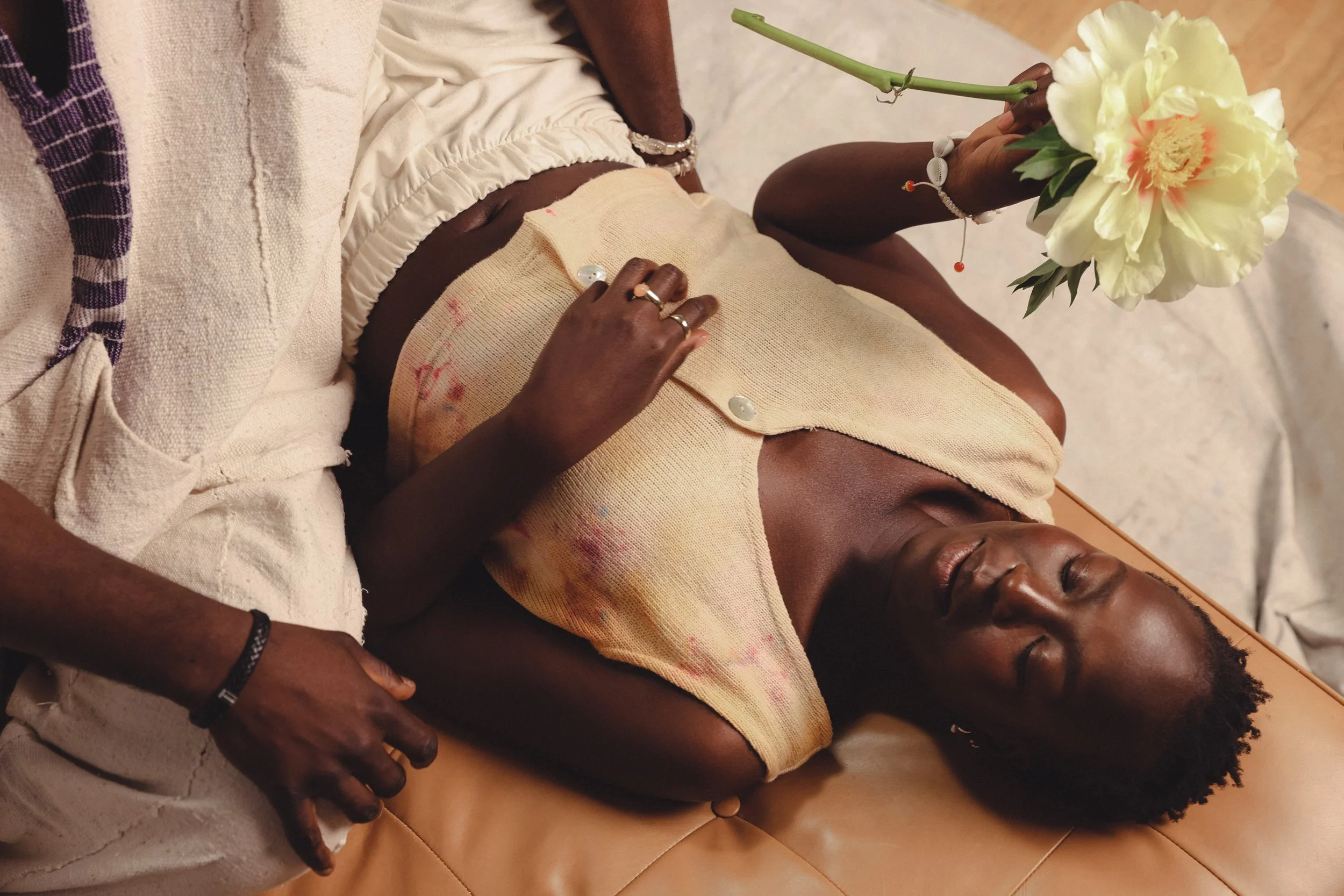 A woman lying on a brown leather surface with eyes closed, holding a bouquet of cream-colored flowers. She wears a sleeveless, tie-dye shirt and jewelry on her wrist and fingers.