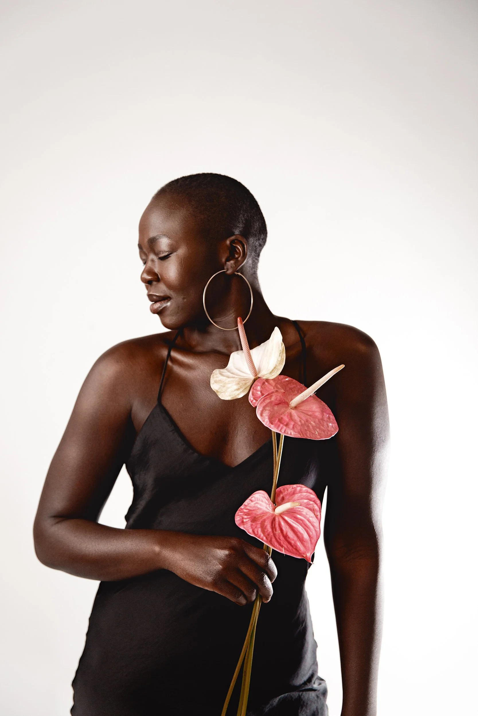 A Black woman with short hair, wearing a black spaghetti strap dress, holding a stem with pink and white Anthurium flowers, and wearing large hoop earrings, is posed against a plain white background.