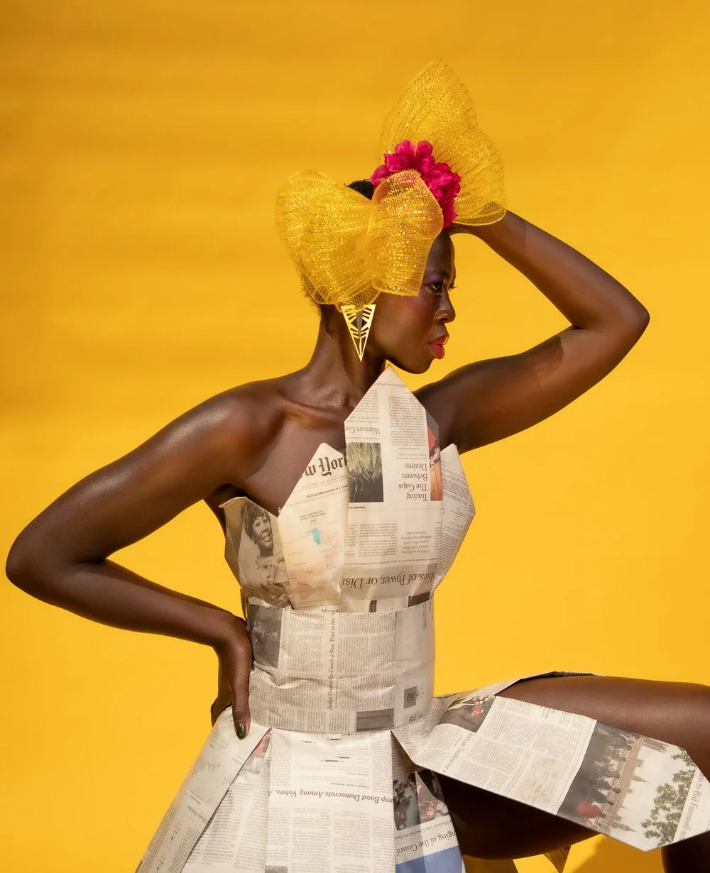 A woman with dark skin wearing a dress made of newspaper pages, posing against a yellow background. She has an elaborate yellow headpiece with pink flowers and large earrings.