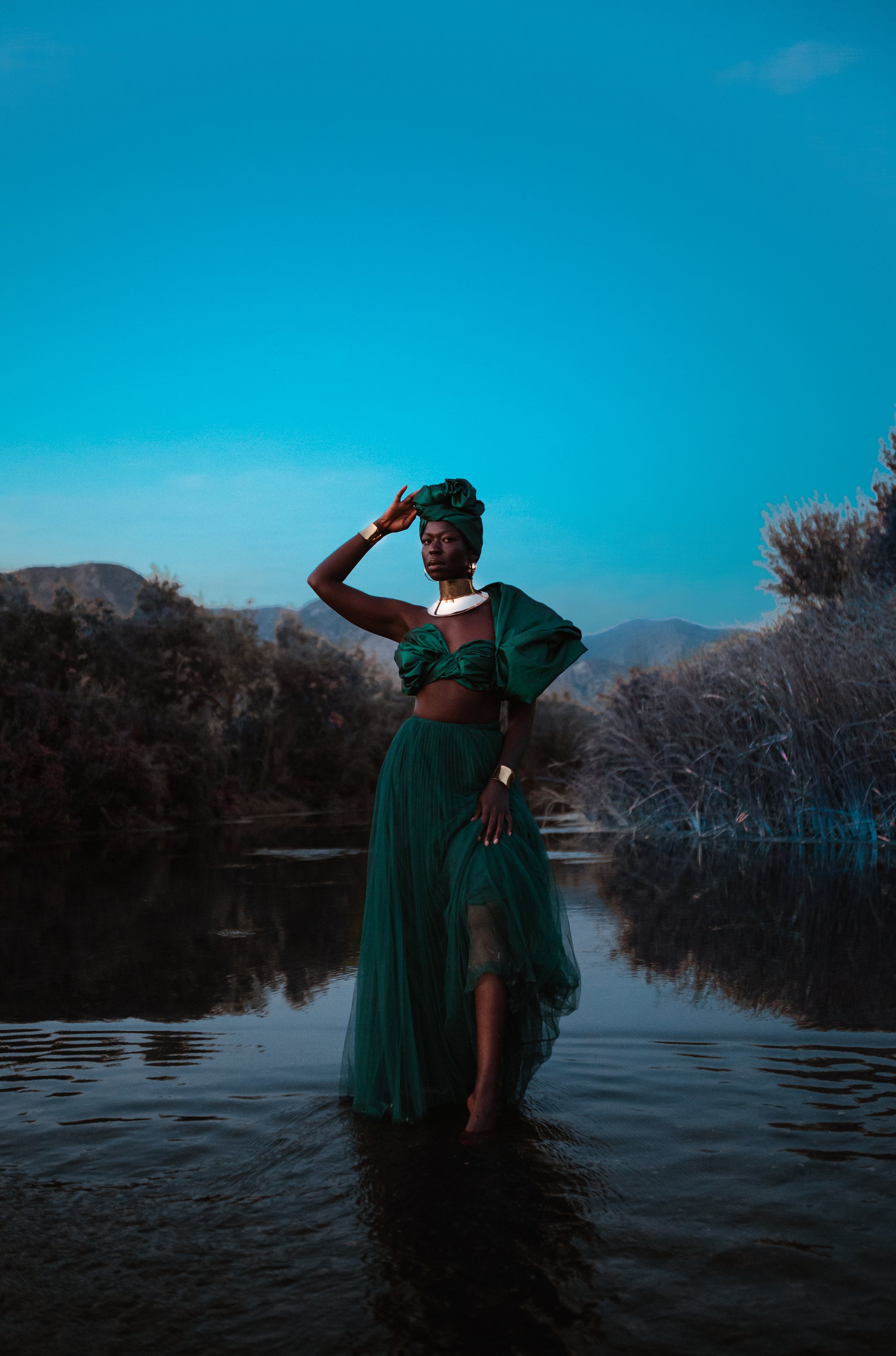 A woman in a green dress and headwrap stands in a river during dusk, with mountains and trees in the background.