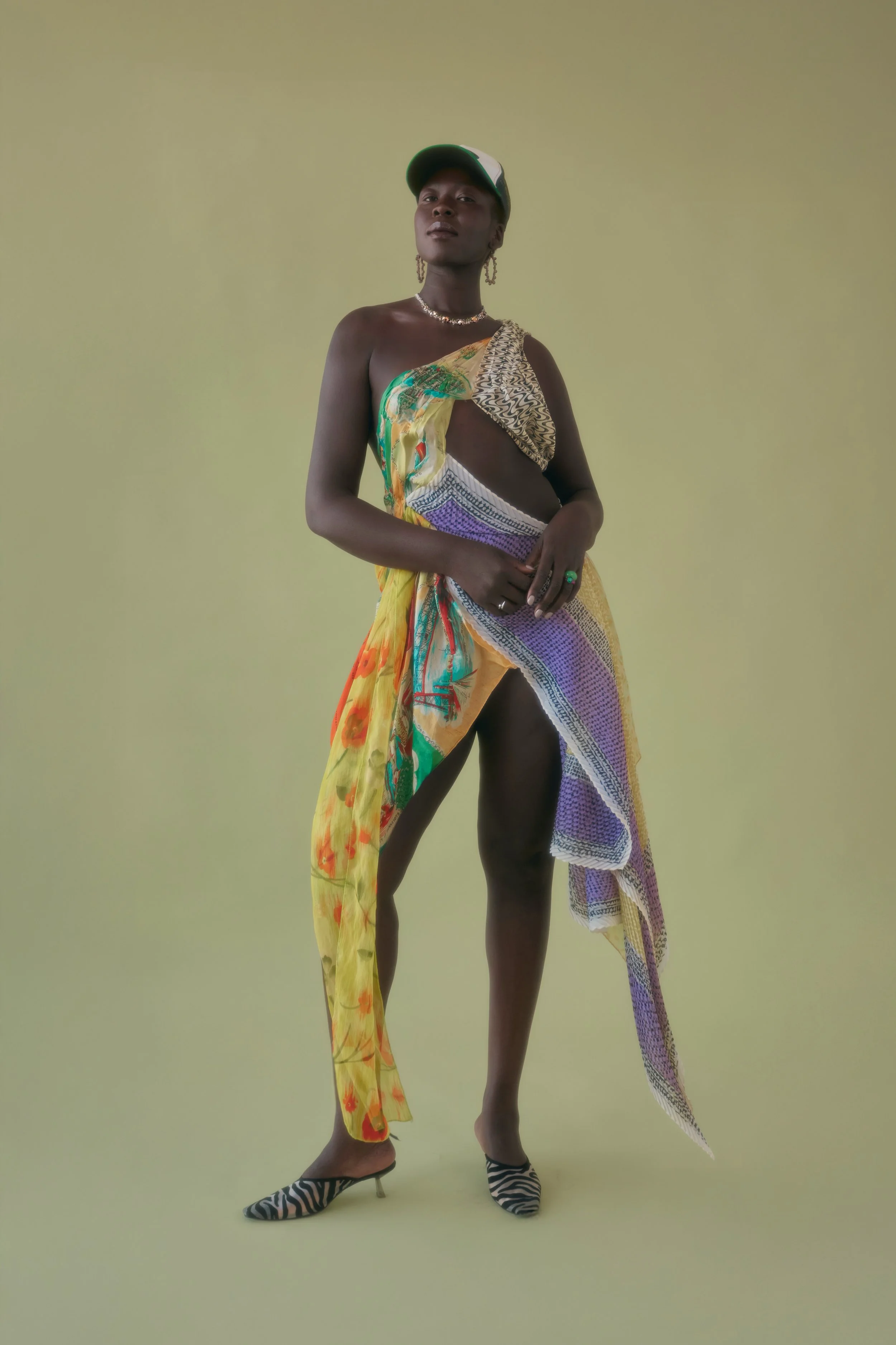 A person standing against a plain background, wearing a colorful tie-dye dress with a slit, zebra-print heels, a baseball cap, and jewelry.