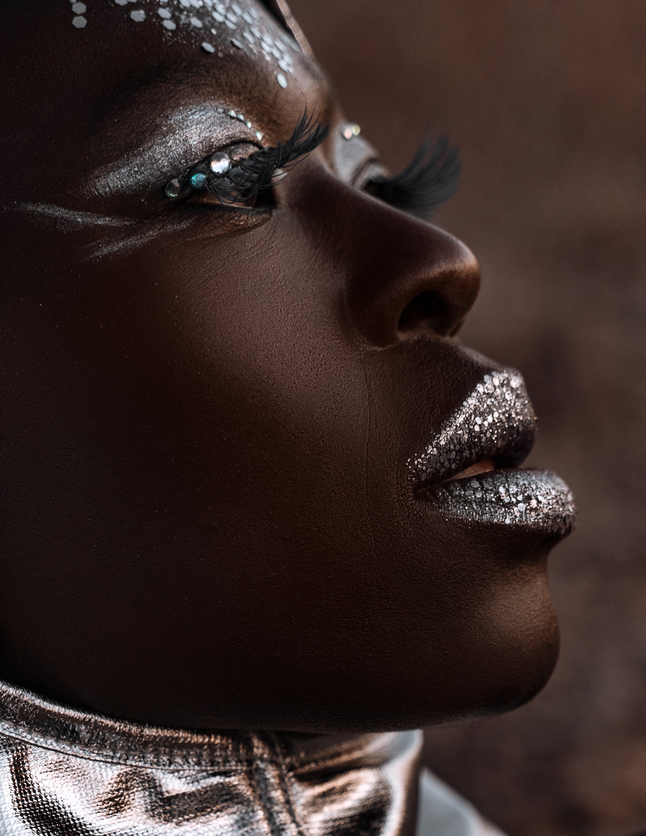 Close-up of a person's face with metallic silver makeup on lips, eyelids, and eyelashes, and adorned with small rhinestones.