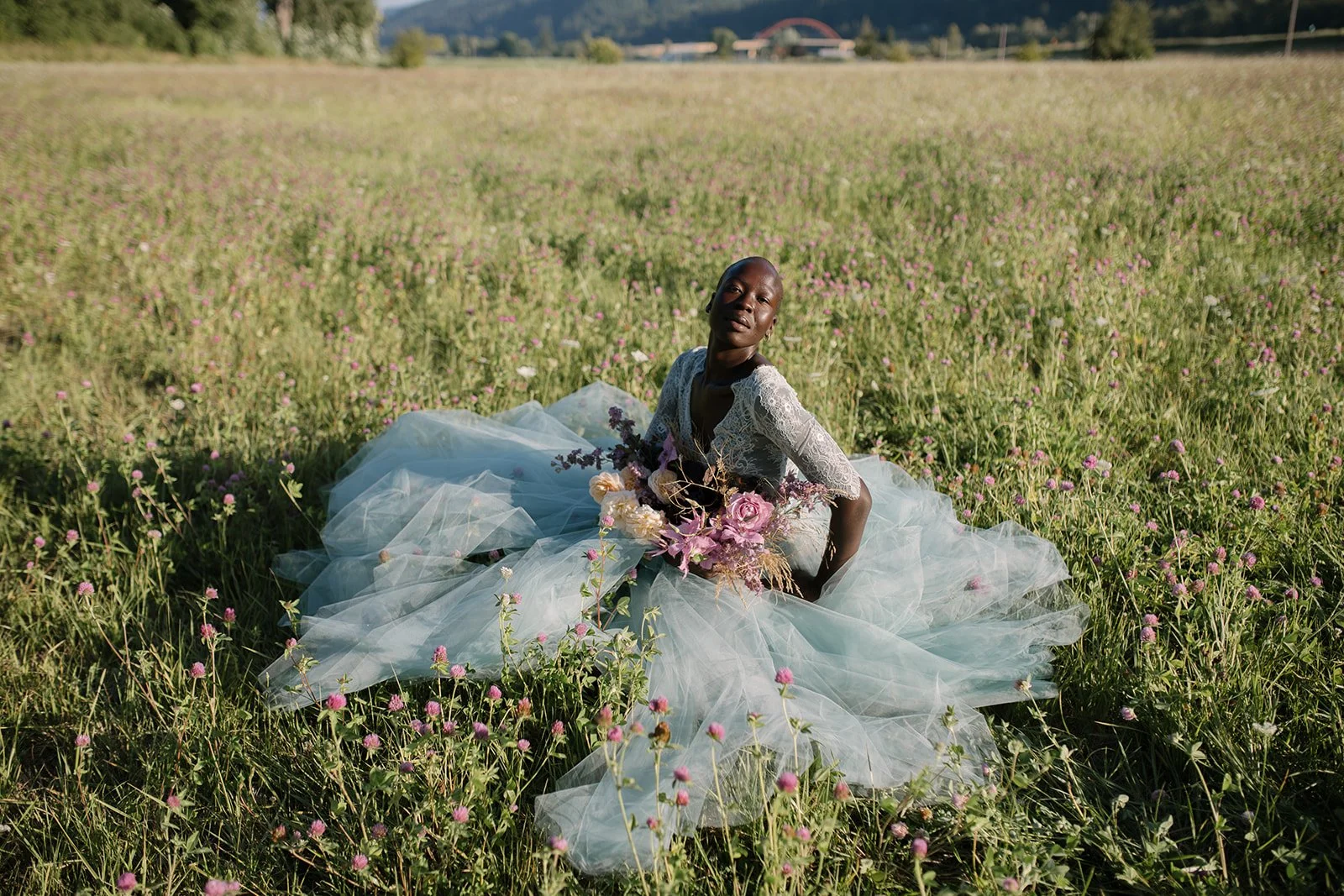 A woman in a white lace dress sitting in a field of pink flowers holding a bouquet.