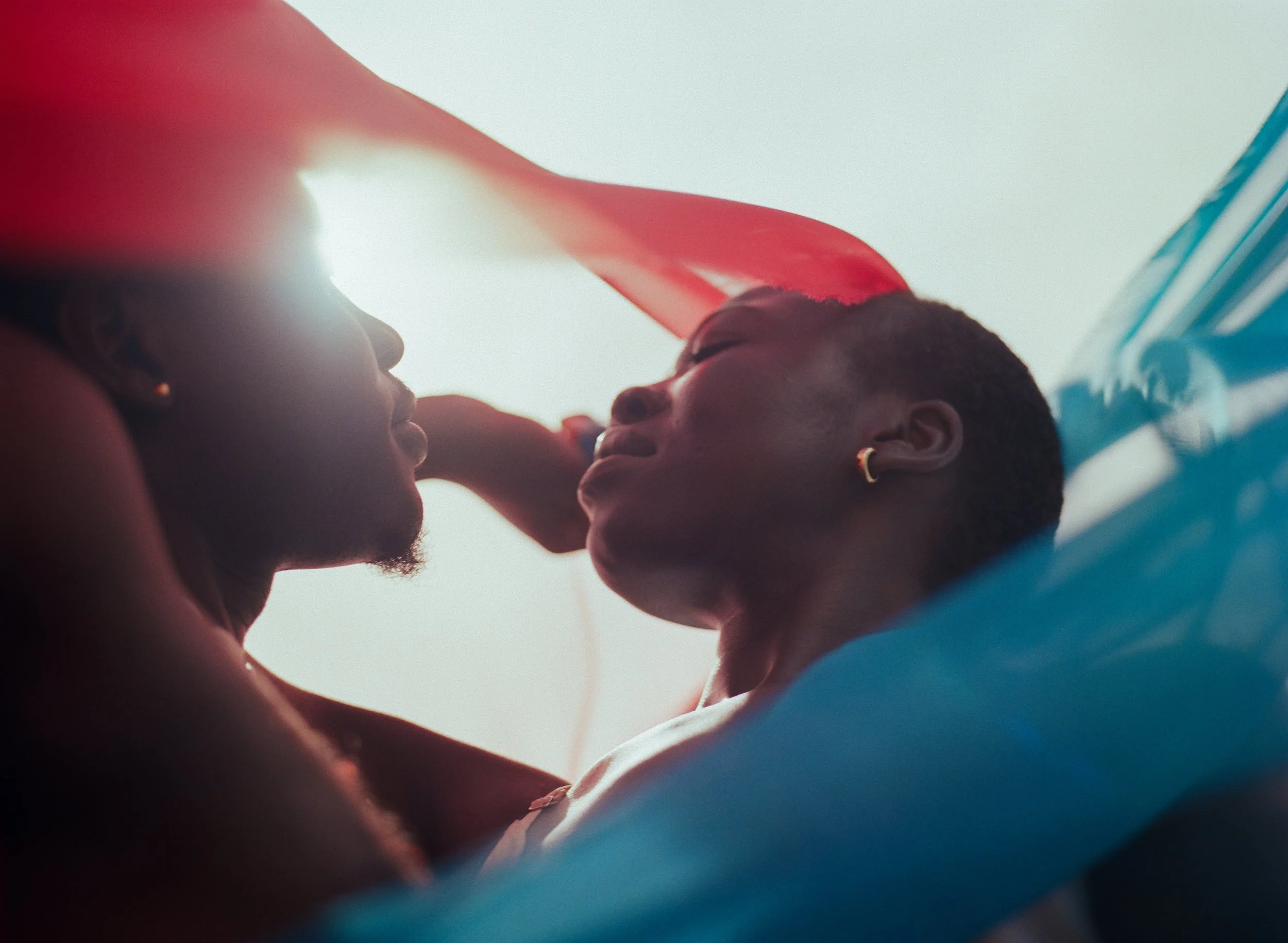 Two women lying close together, possibly on a beach, with a red and blue canopy or fabric above them, sharing an intimate moment.