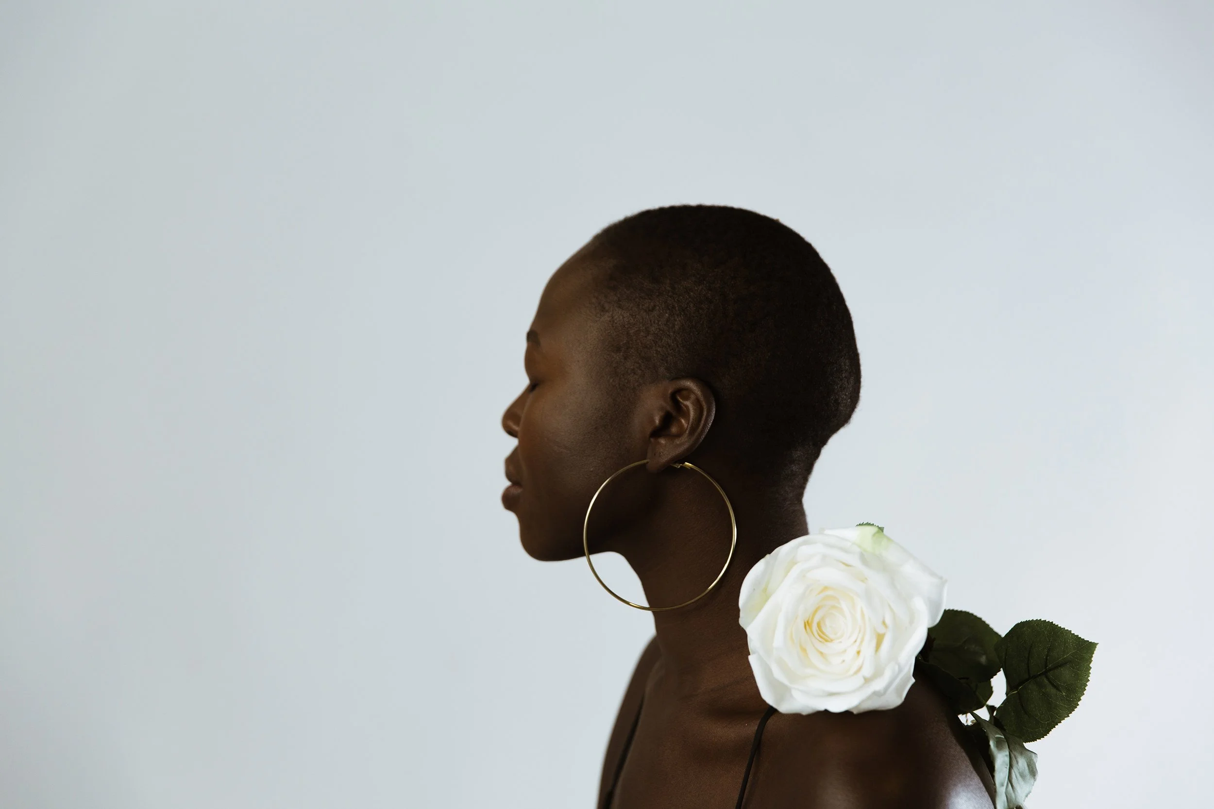 Side profile of a woman with short hair wearing large hoop earrings, a white rose on her shoulder, and a dark top, against a plain light background.