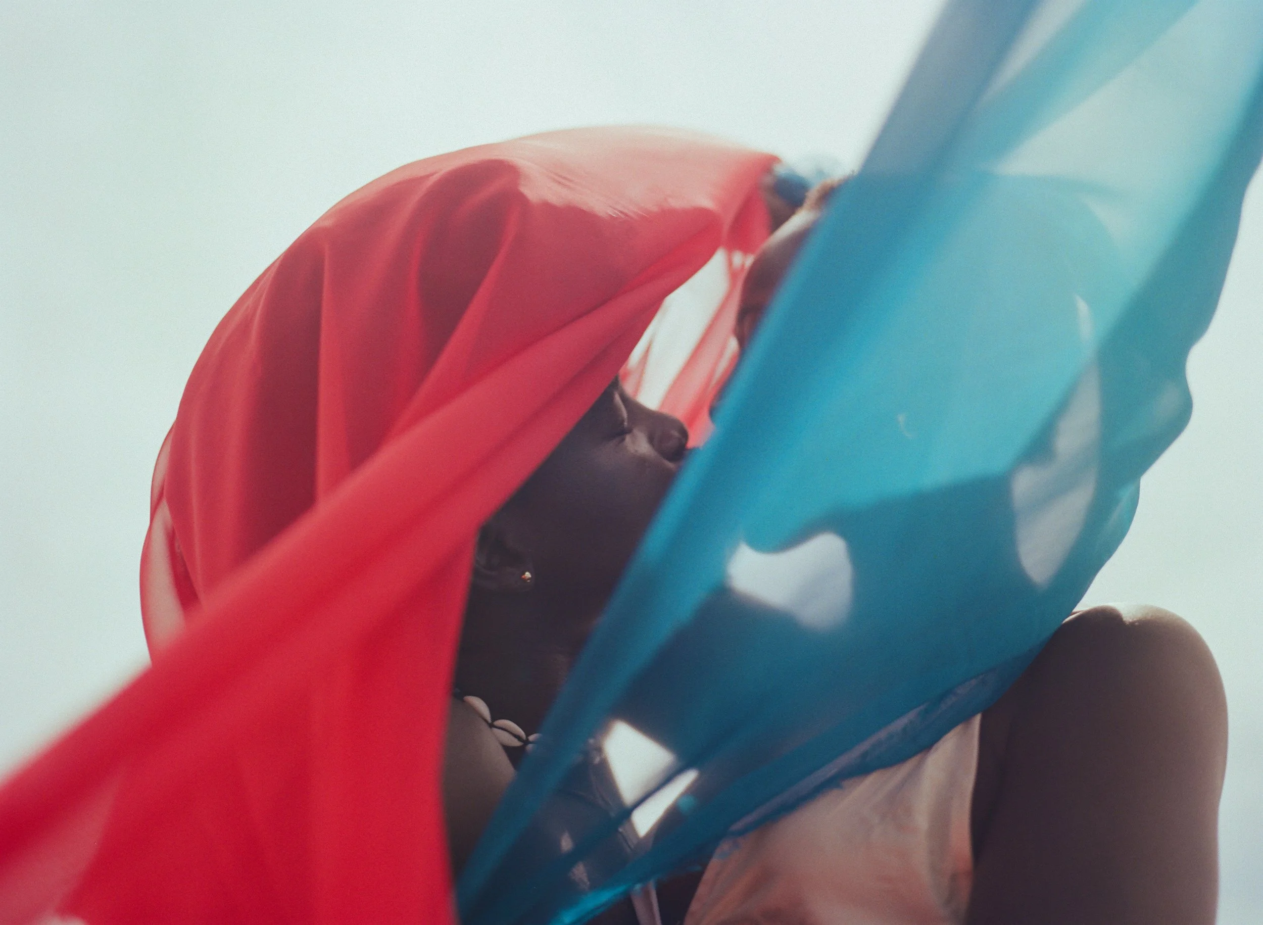 A person draped in vibrant red and orange fabric, holding a translucent blue object, taken from a low angle with a clear sky in the background.
