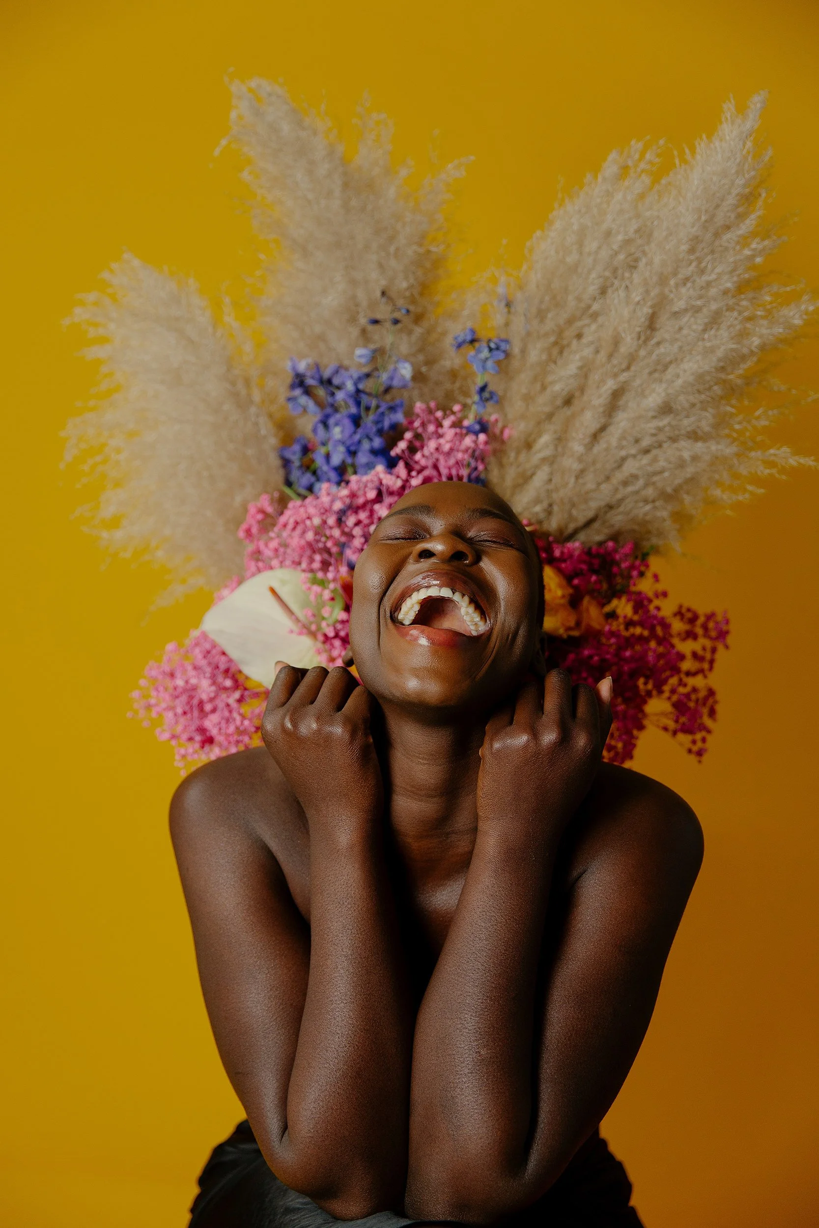 A woman laughing joyfully with eyes closed, surrounded by colorful flowers and dried pampas grass against a yellow background.