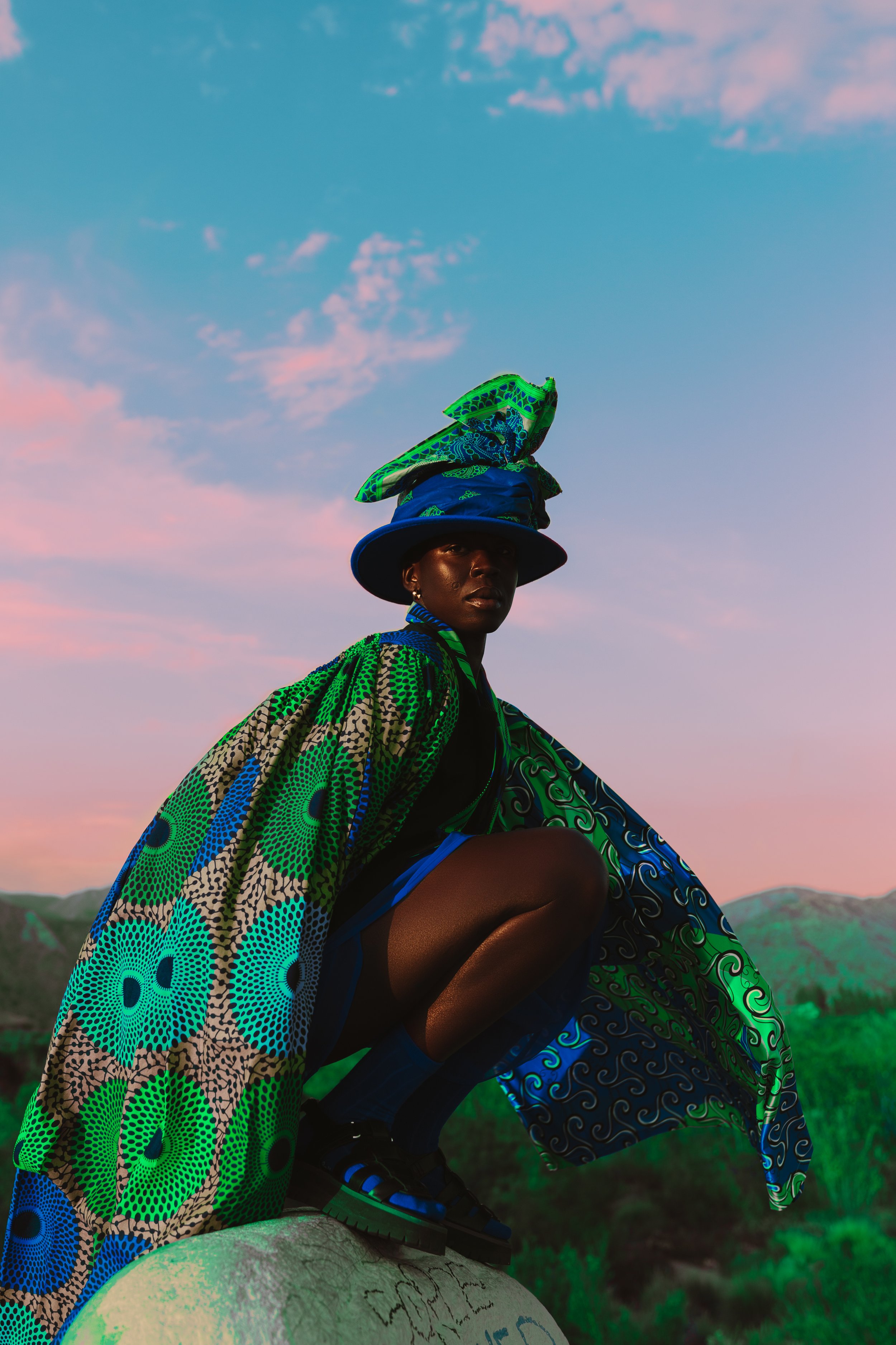 Person wearing colorful patterned clothing and a hat with a bird design, squatting on a large stone outdoors, with hills and a colorful sky in the background.