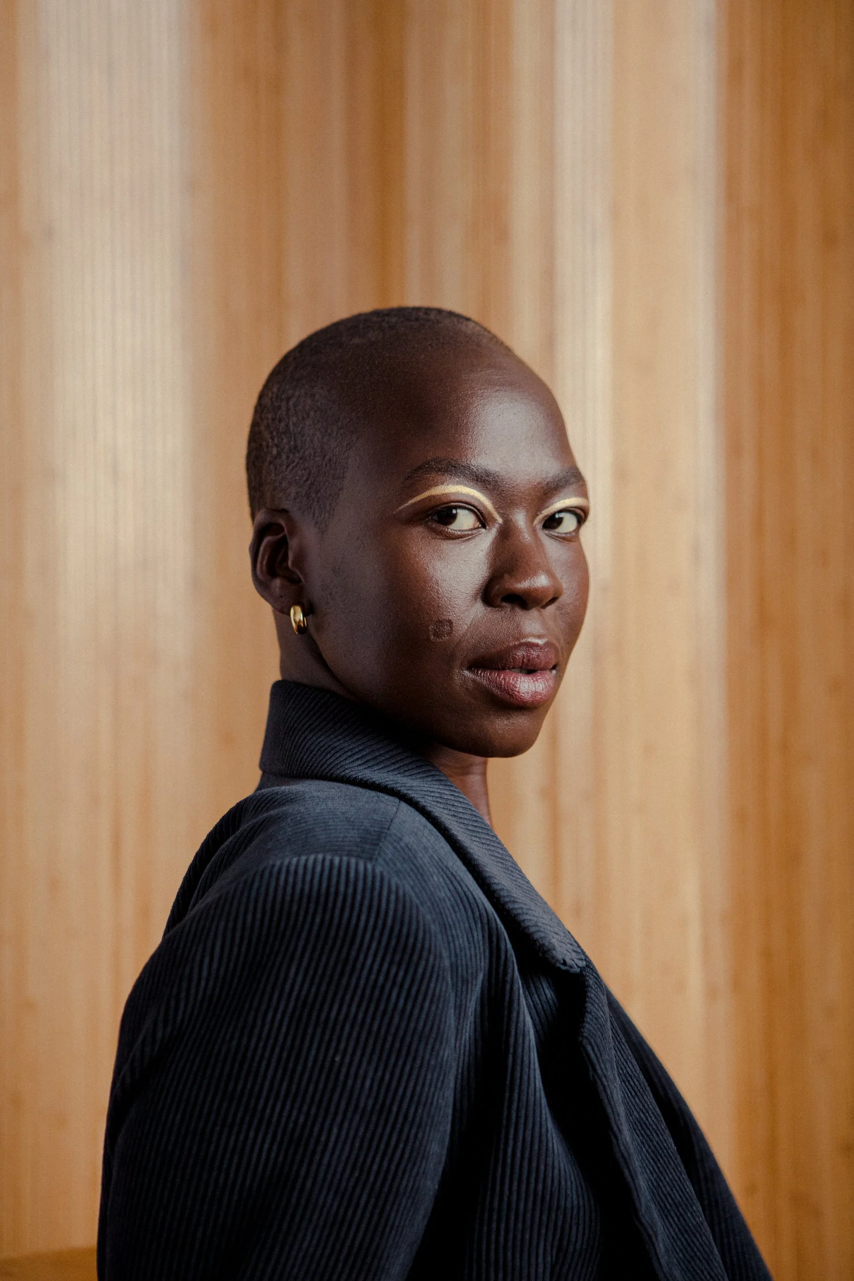 A portrait of a Black woman with a shaved head, wearing a dark blazer, gold earrings, and standing in front of a wooden background.