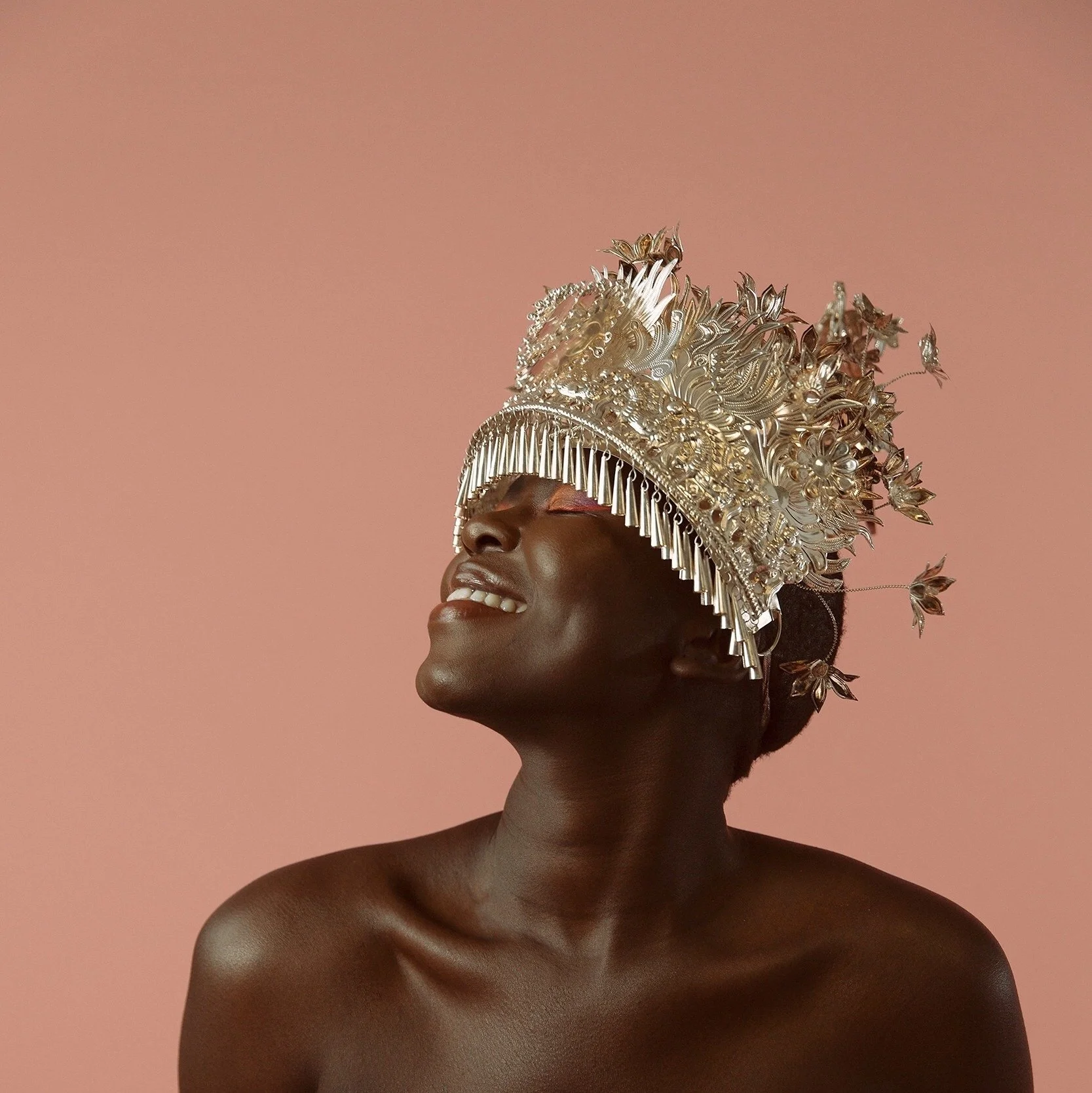 A woman with dark skin and short hair wearing an ornate silver crown adorned with intricate patterns, flowers, and leaves, posing against a pink background.