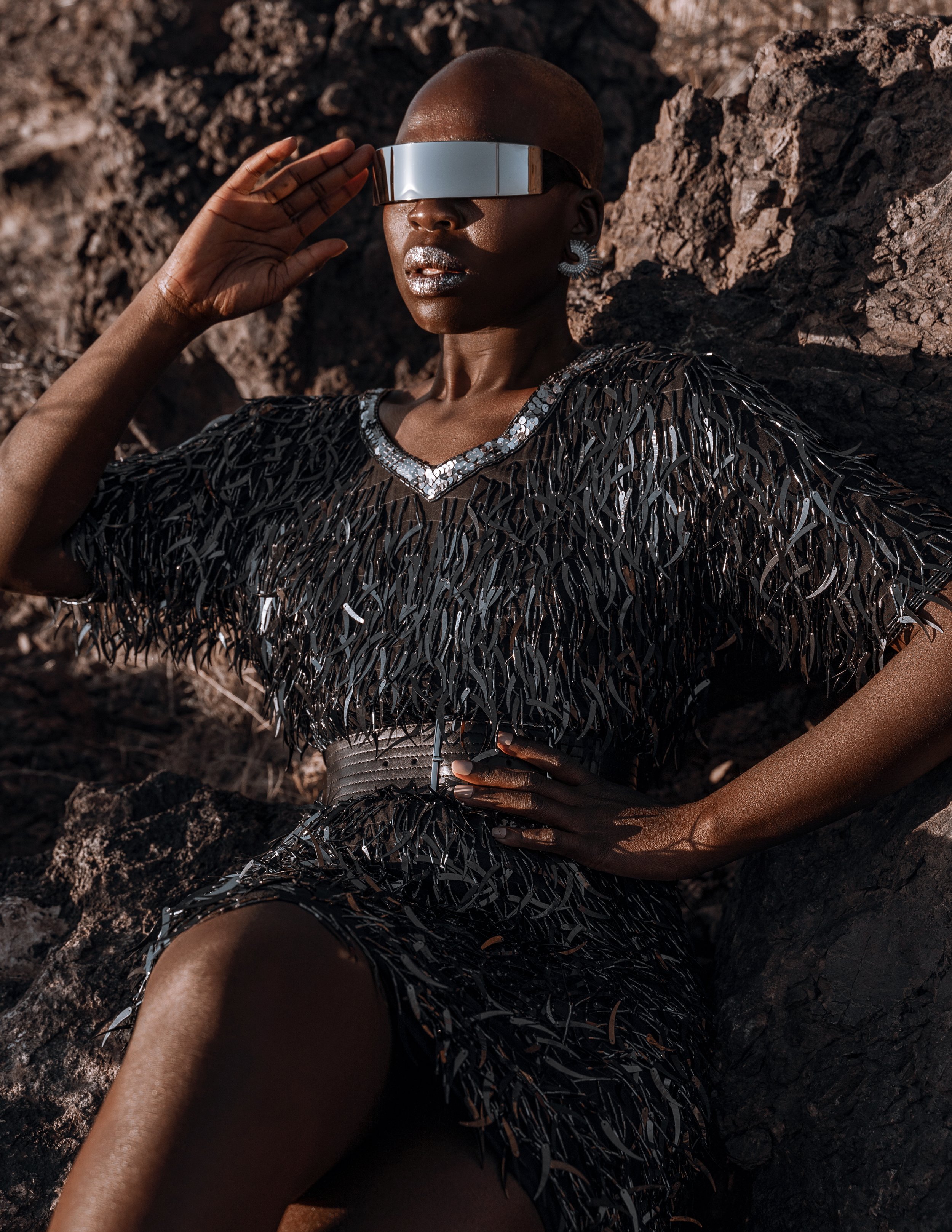 A woman with dark skin wearing futuristic sunglasses, a black feathered dress, and large earrings, sitting against rocks outdoors.