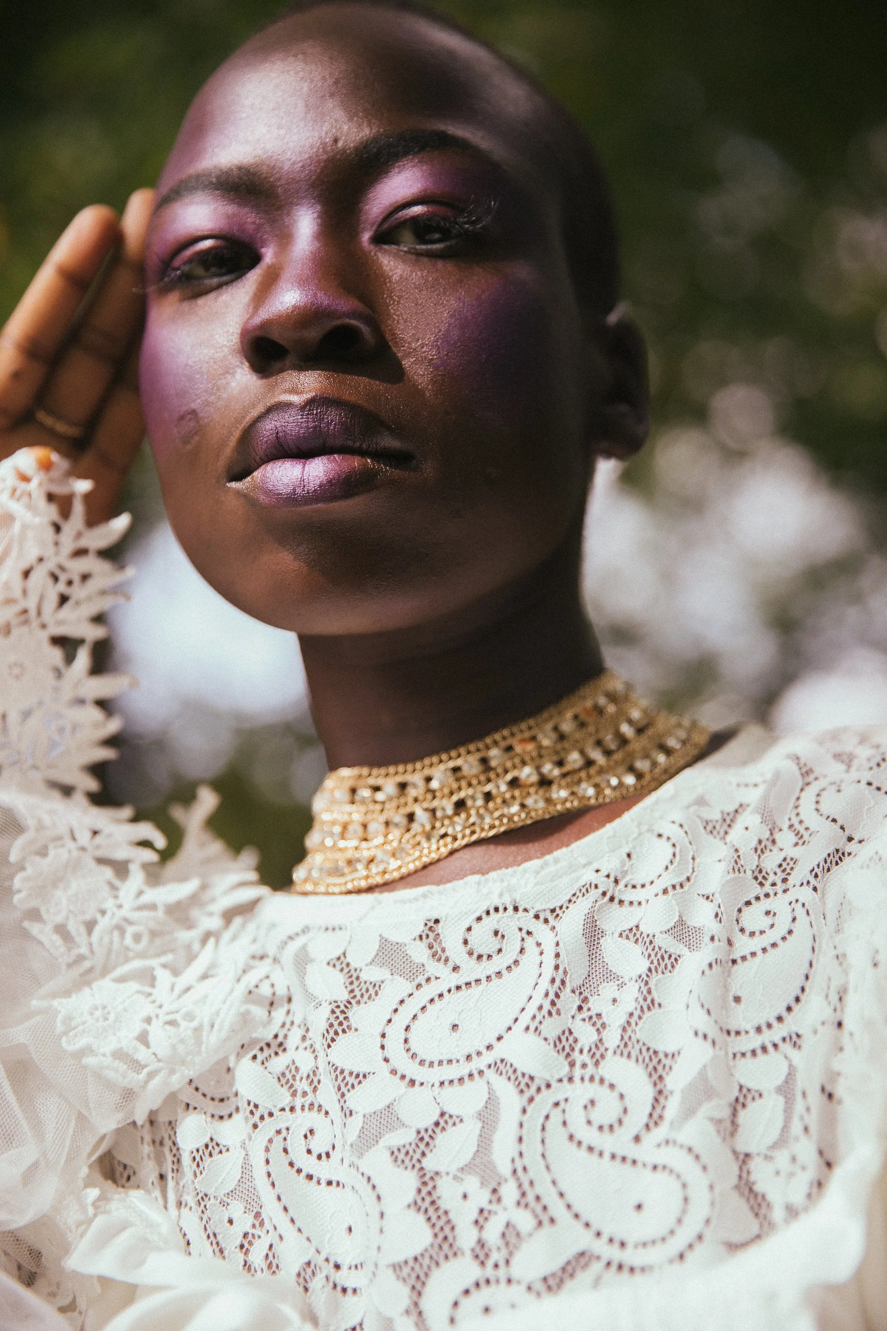 A close-up of a woman with dark skin and short hair, wearing a white lace dress and layered gold necklace, posing outdoors.