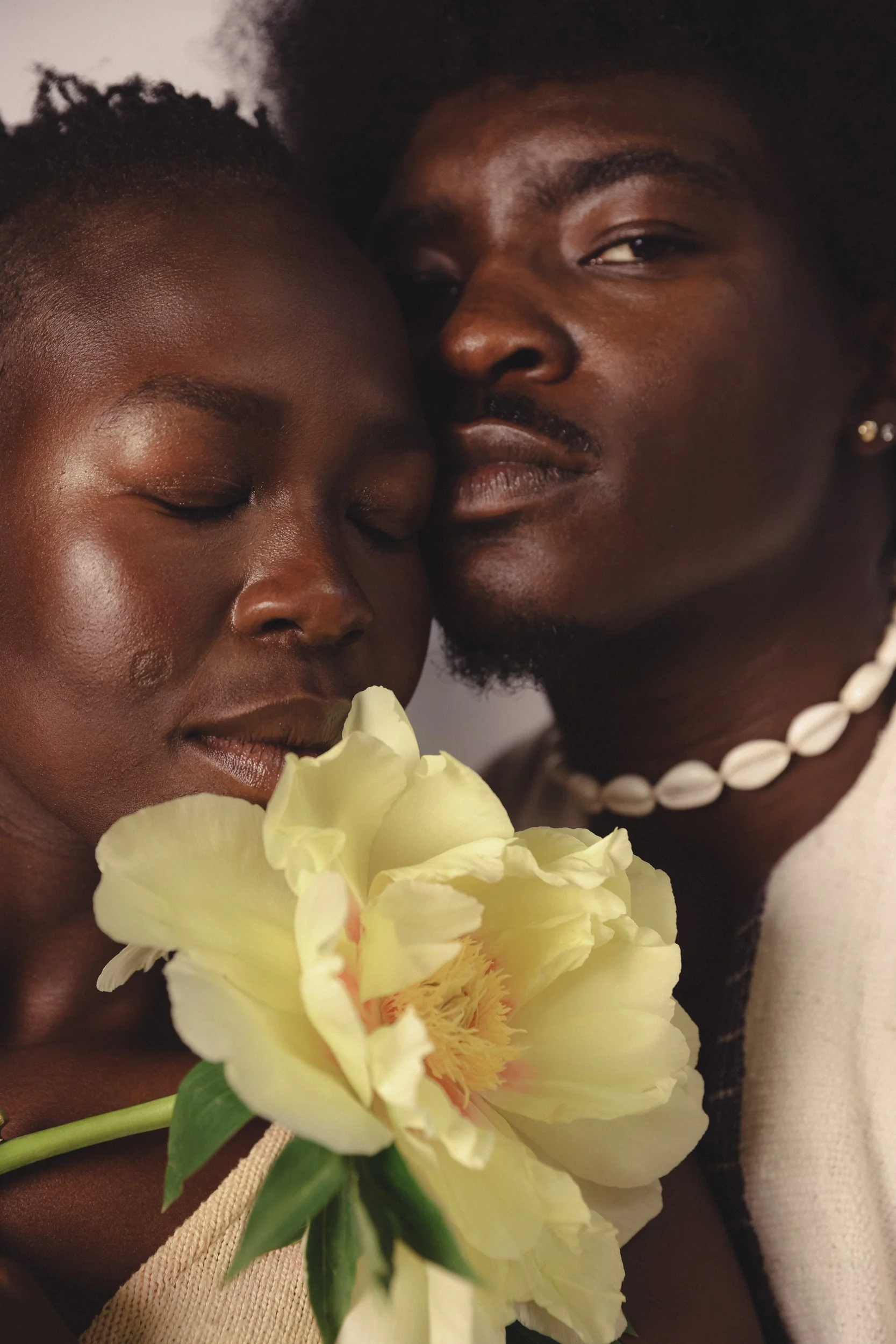 Close-up of a smiling woman with eyes closed holding a large pale yellow flower, and a man with dark skin and black hair close to her, both with serene expressions.