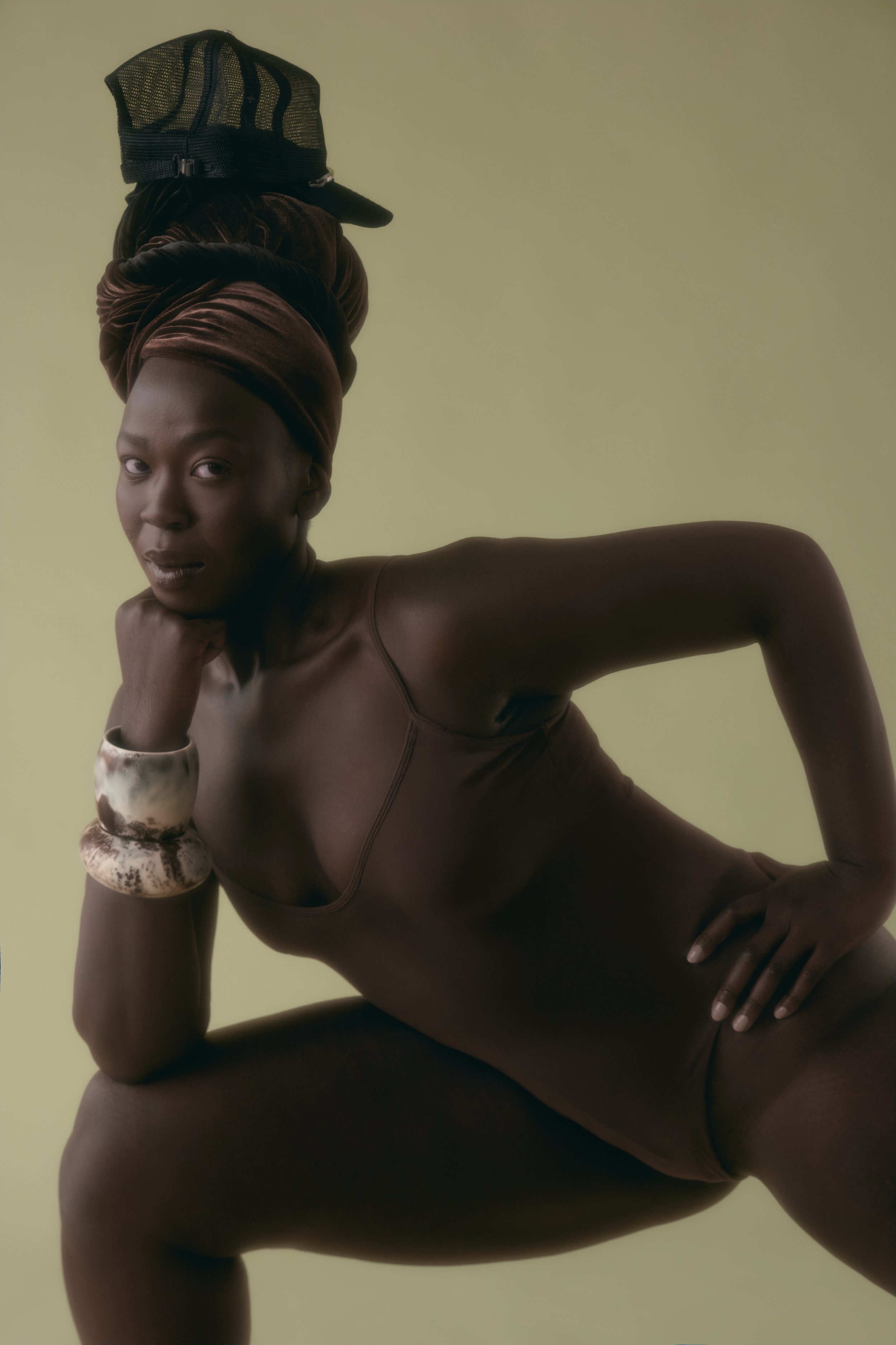 A woman with dark skin, wearing a head wrap and jewelry, poses against a plain background with one hand on her chin and the other on her hip.