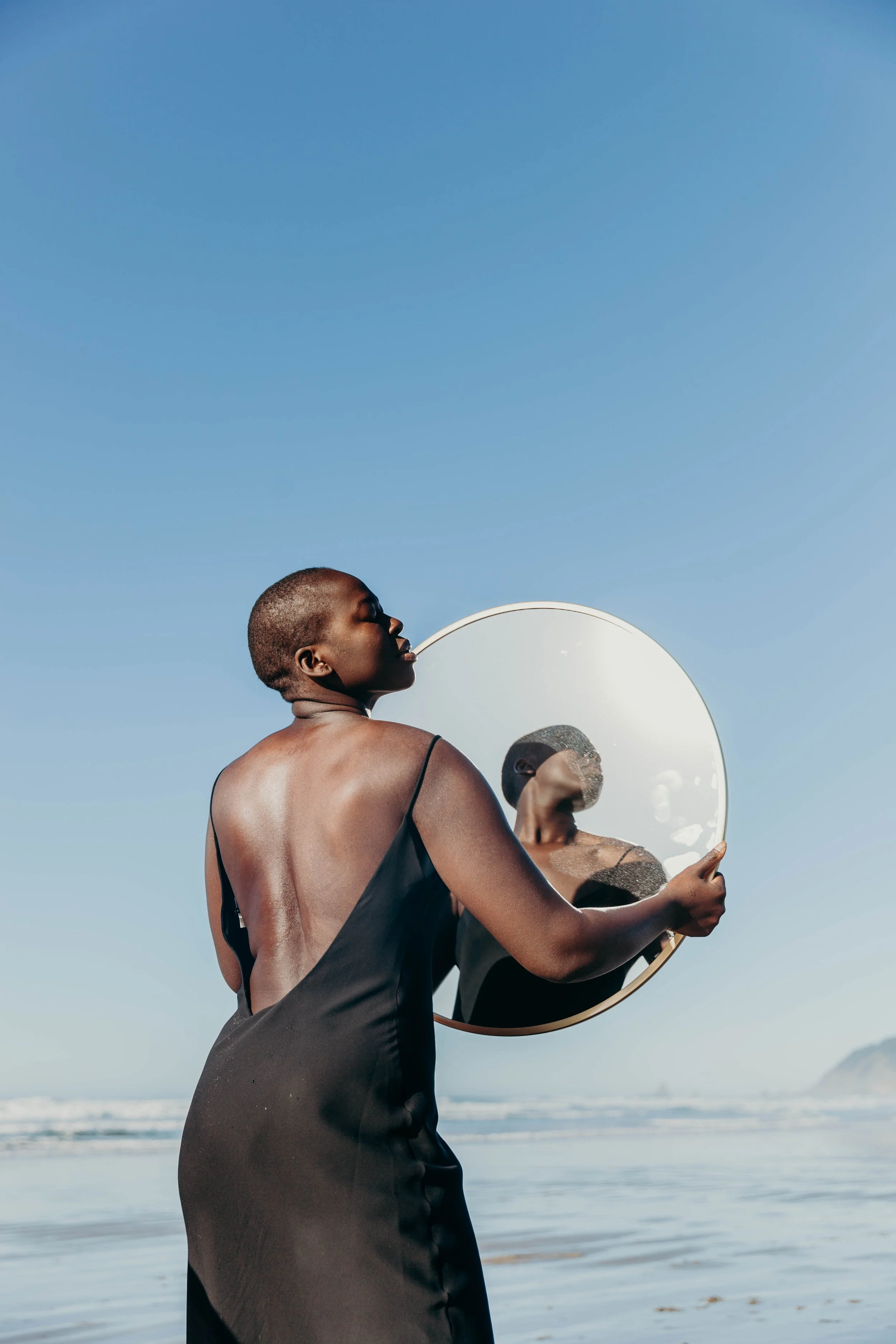 A woman in a black dress holding a round mirror on a beach under a clear sky.