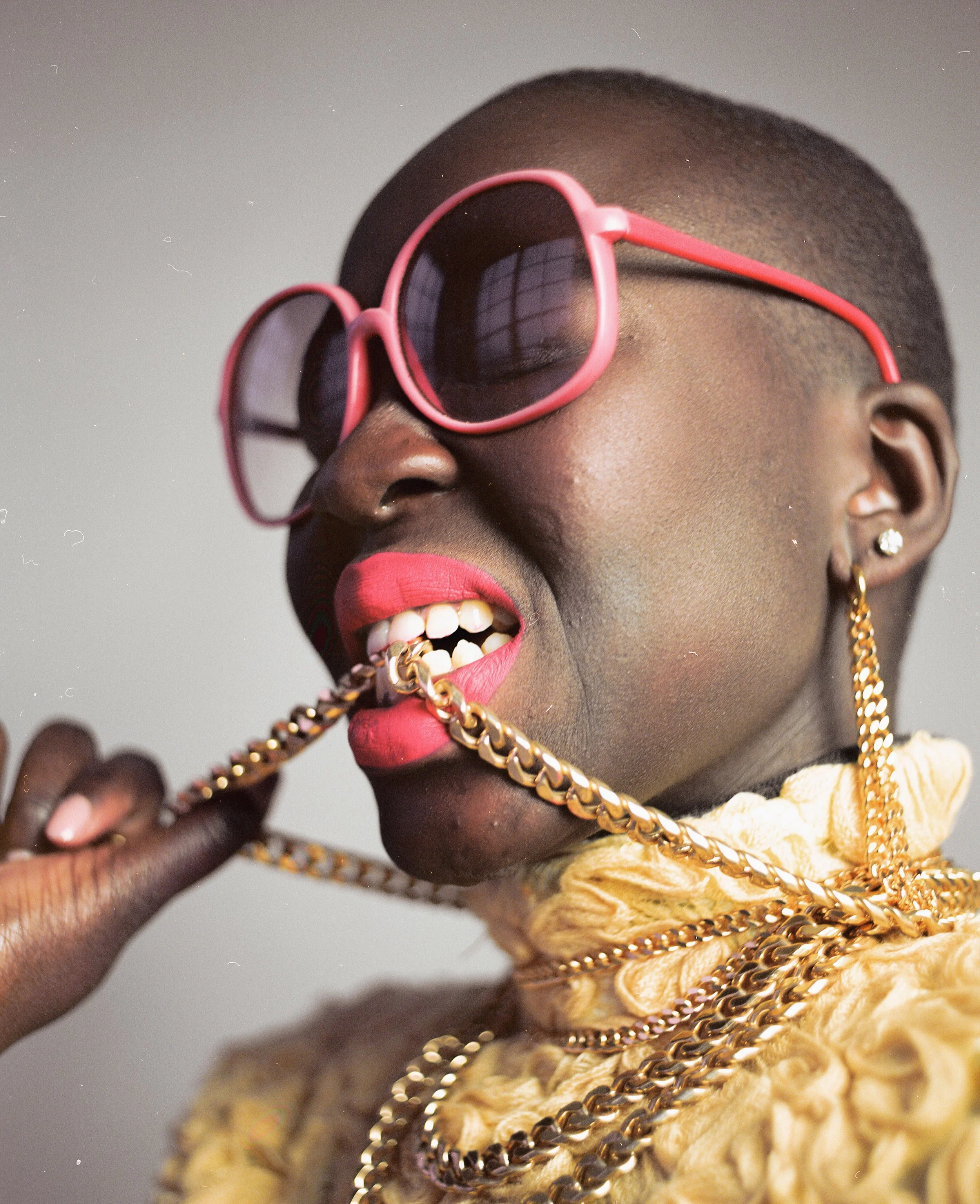 A person wearing pink sunglasses with dark lenses, red lipstick, and gold jewelry, holding a gold chain in their mouth.