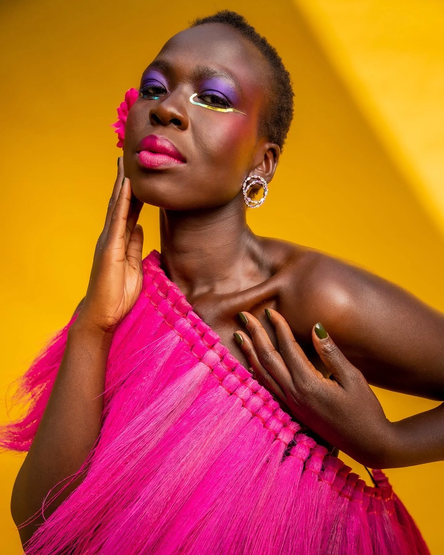 Portrait of a woman with dark skin, wearing pink and purple makeup, a pink dress, and gold earrings, with a bright yellow background.