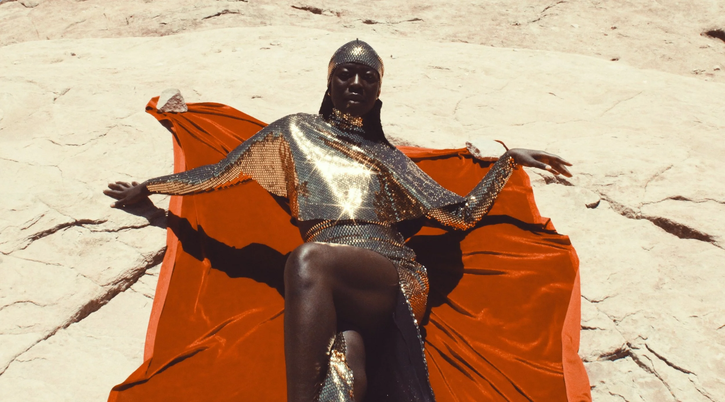 A person dressed in a shiny, sequined outfit, lying on an orange fabric spread on cracked, dry ground. The person is looking at the camera, with their arms extended and one leg bent.