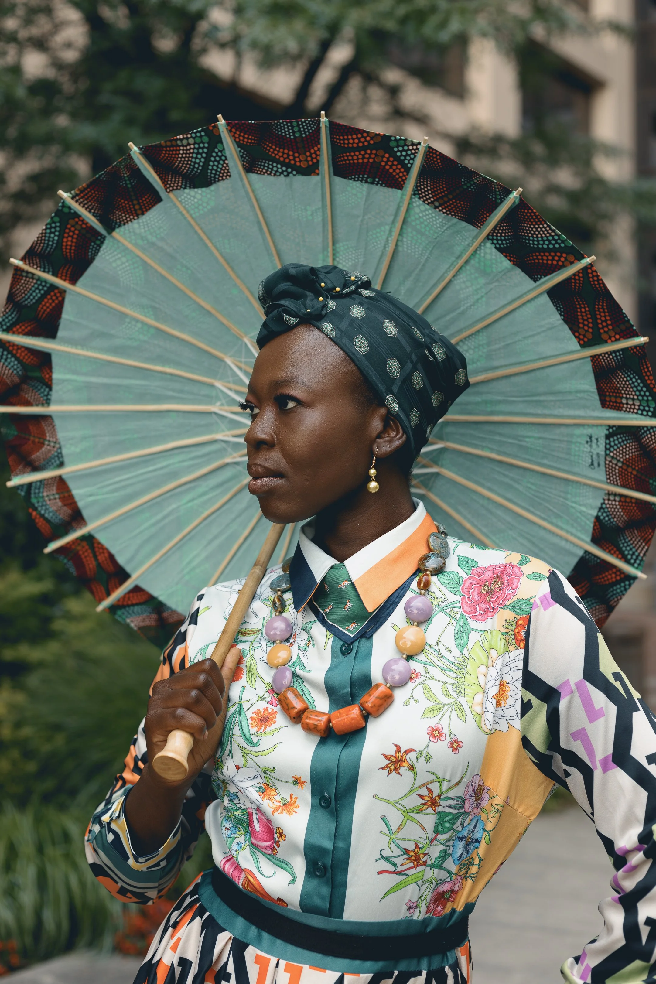 A woman with dark skin wearing a colorful floral top, large statement necklace, and a headscarf, holding an ornate parasol with a patterned border.
