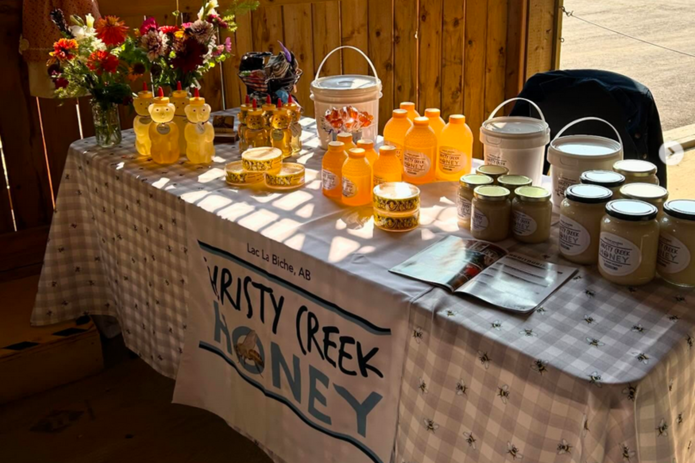 Table displays honey jars, bottles, and beeswax products, with a vase of colorful flowers on the left, set against a wood-paneled background.