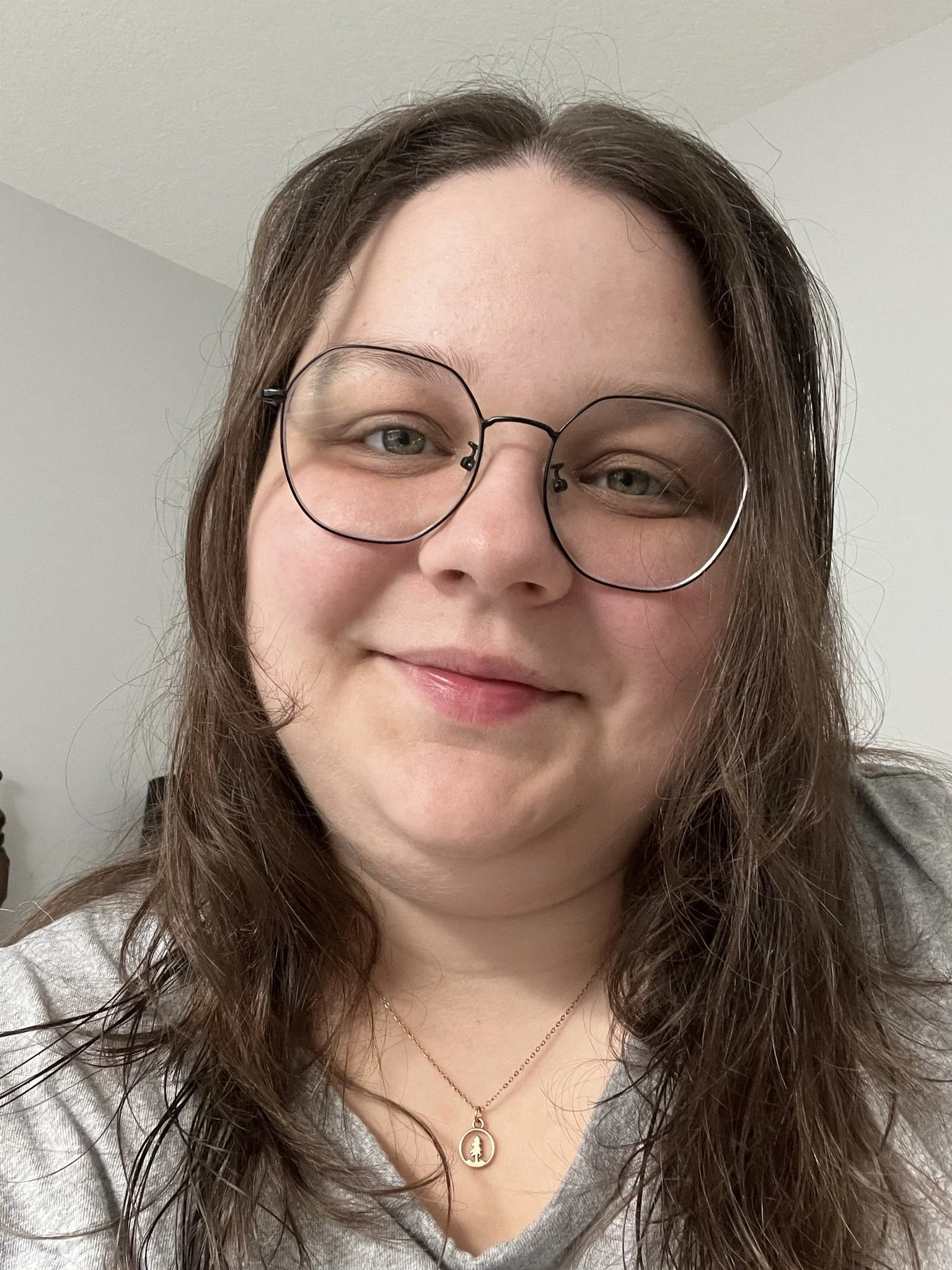 A woman with glasses and wavy brown hair smiling at the camera, wearing a gold necklace with a pendant, in a simple indoor setting.