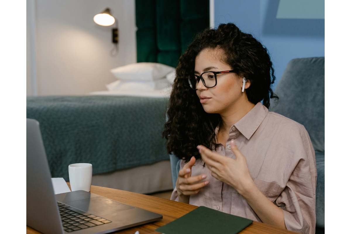 A woman with curly hair, glasses, and AirPods is sitting at a desk in a bedroom, engaging in a video call on her laptop, with a notebook and a white mug nearby.