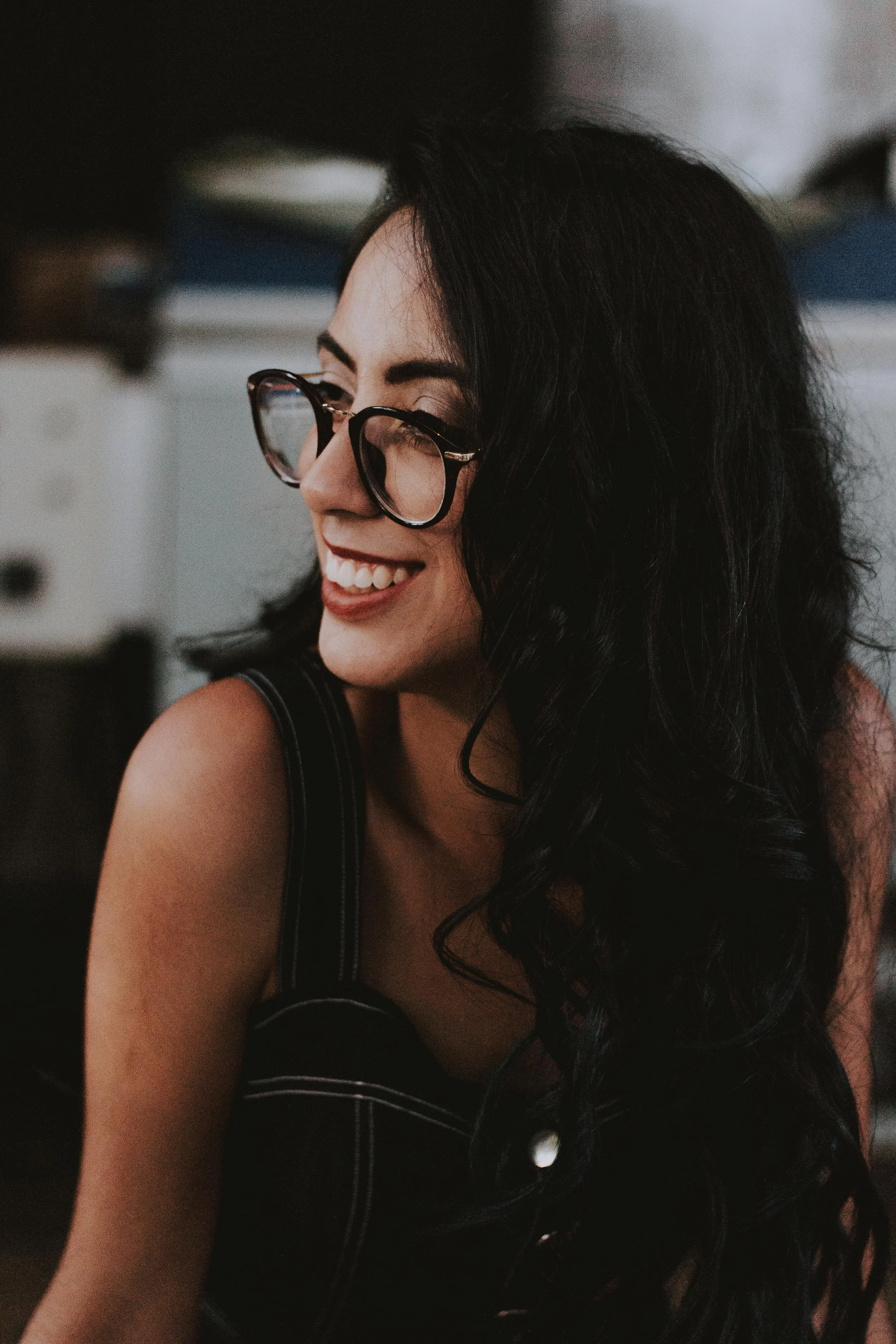 A woman with glasses and dark hair sitting at a wooden table, speaking to someone off-camera, with a laptop, croissants, coffee cups, and a vase with purple flowers in a bright, loft-style room with brick walls and large windows.