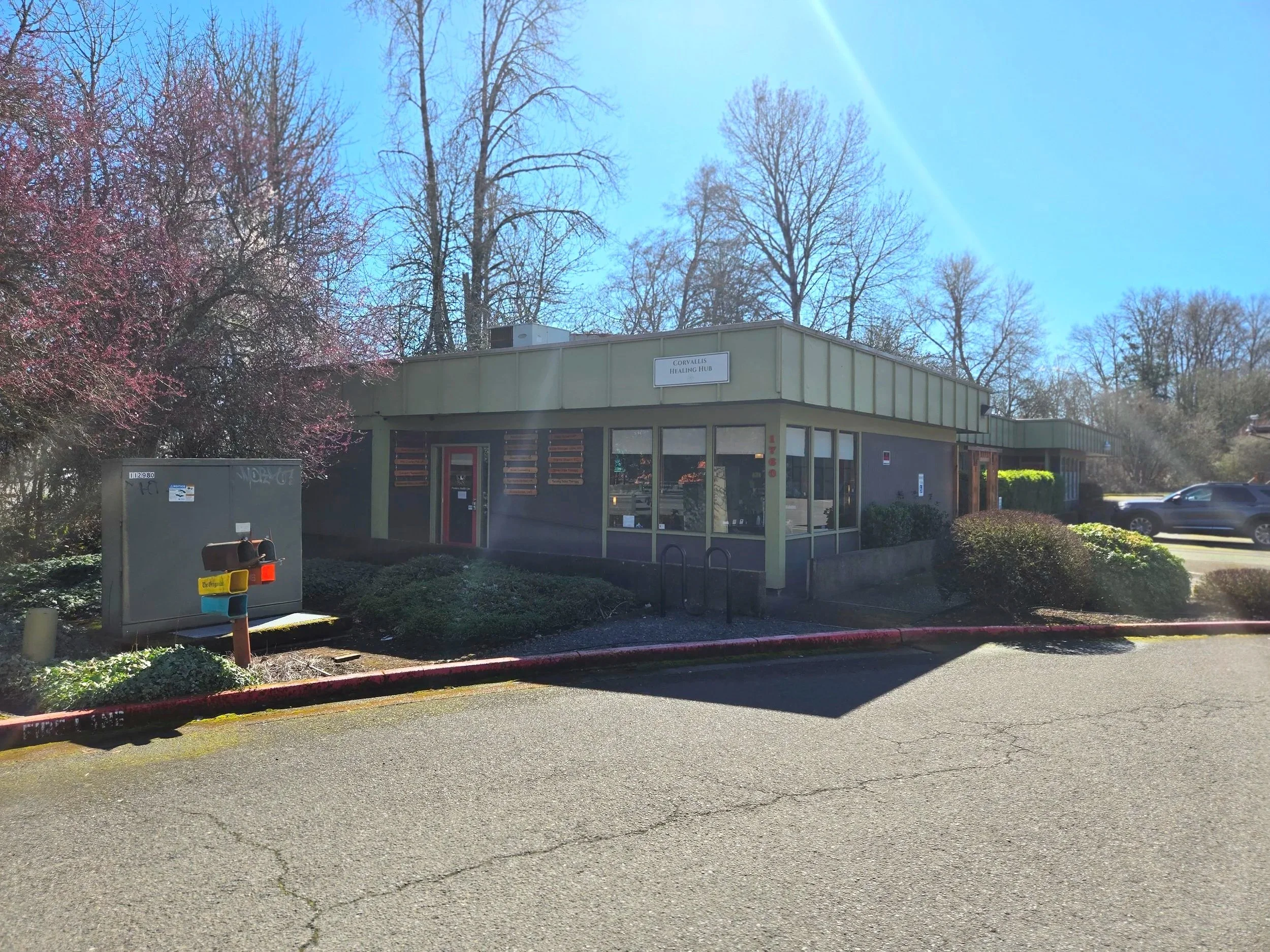 A building with a sign that reads 'Corvallis Healing Hub', surrounded by bushes and trees, with parked cars on the side and a clear blue sky above.
