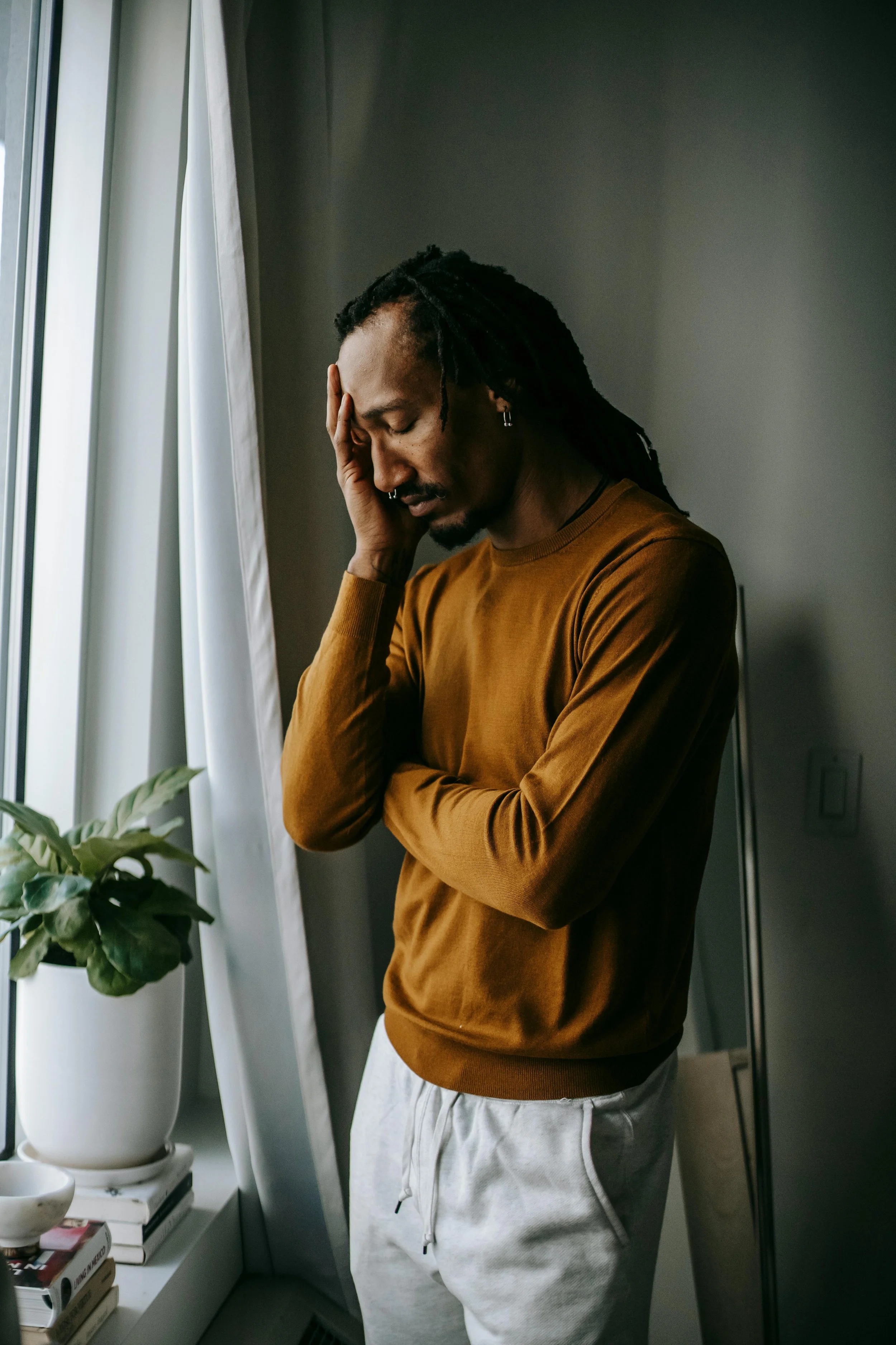 A man with dreadlocks and earrings, wearing a brown sweatshirt and gray sweatpants, stands by a window with a potted plant and books nearby. He appears distressed, with one hand on his forehead and his eyes closed.