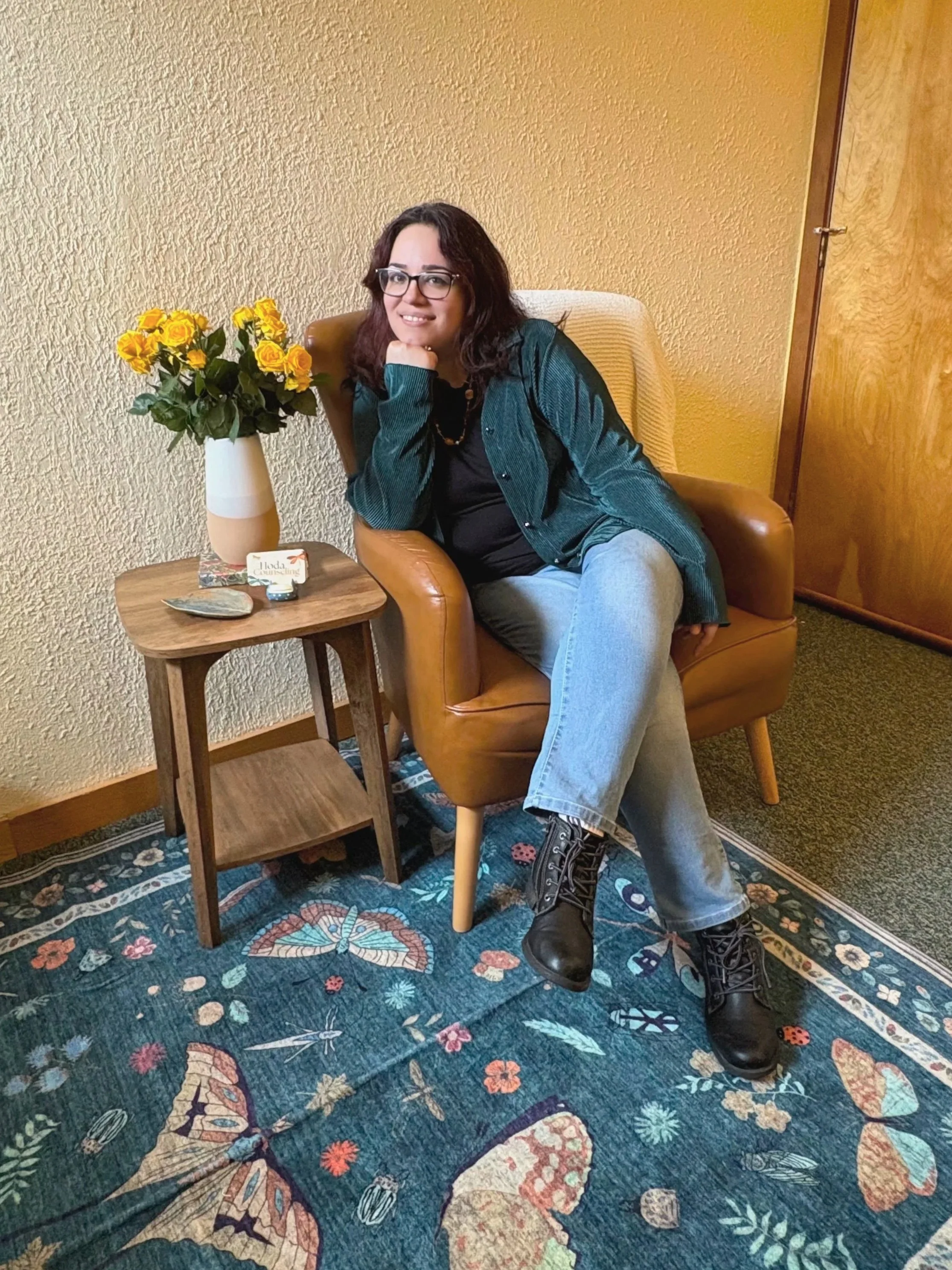 A woman with glasses and dark hair sitting on a tan armchair, smiling, wearing a dark green jacket, black top, and gray jeans. She is sitting next to a small wooden side table with a white vase of yellow roses and some small items. The floor has a decorative blue rug with butterfly and floral patterns.