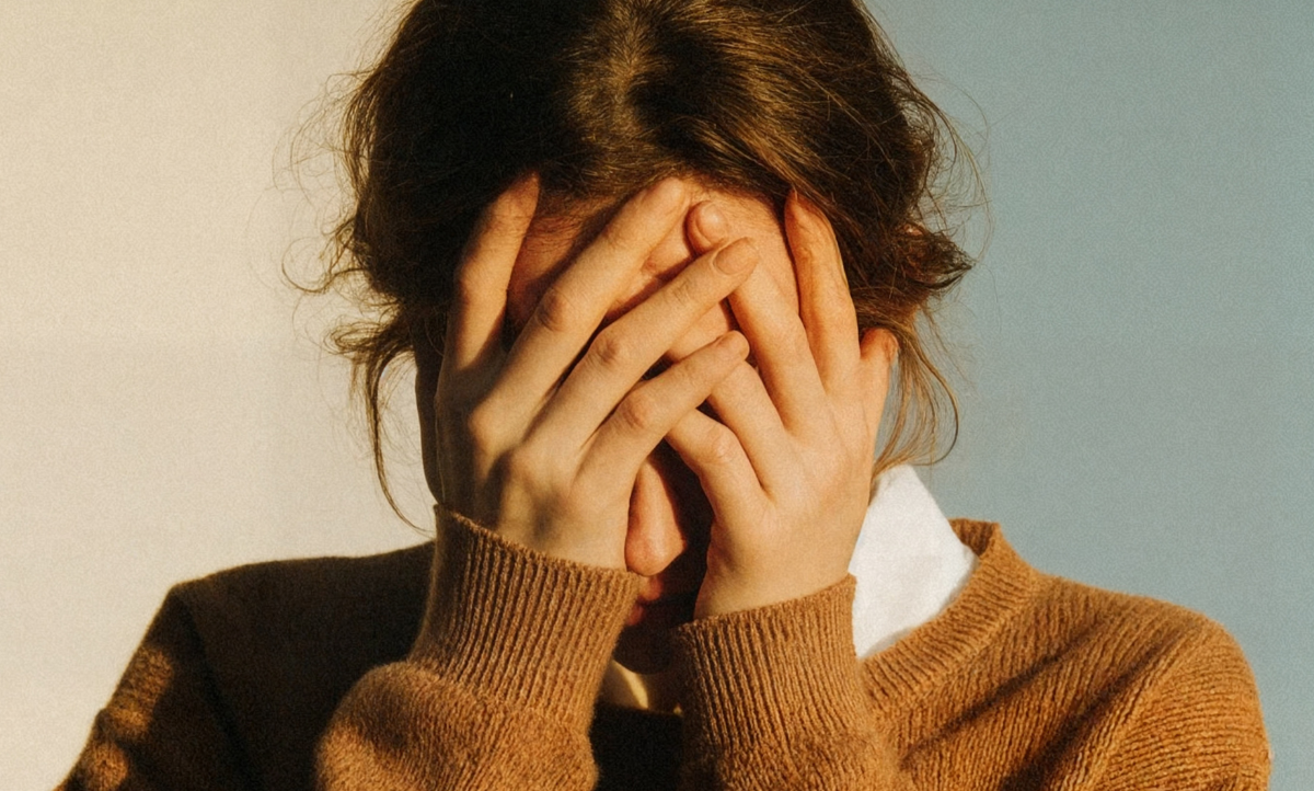 Person with brown hair covering face with hands, wearing a brown sweater