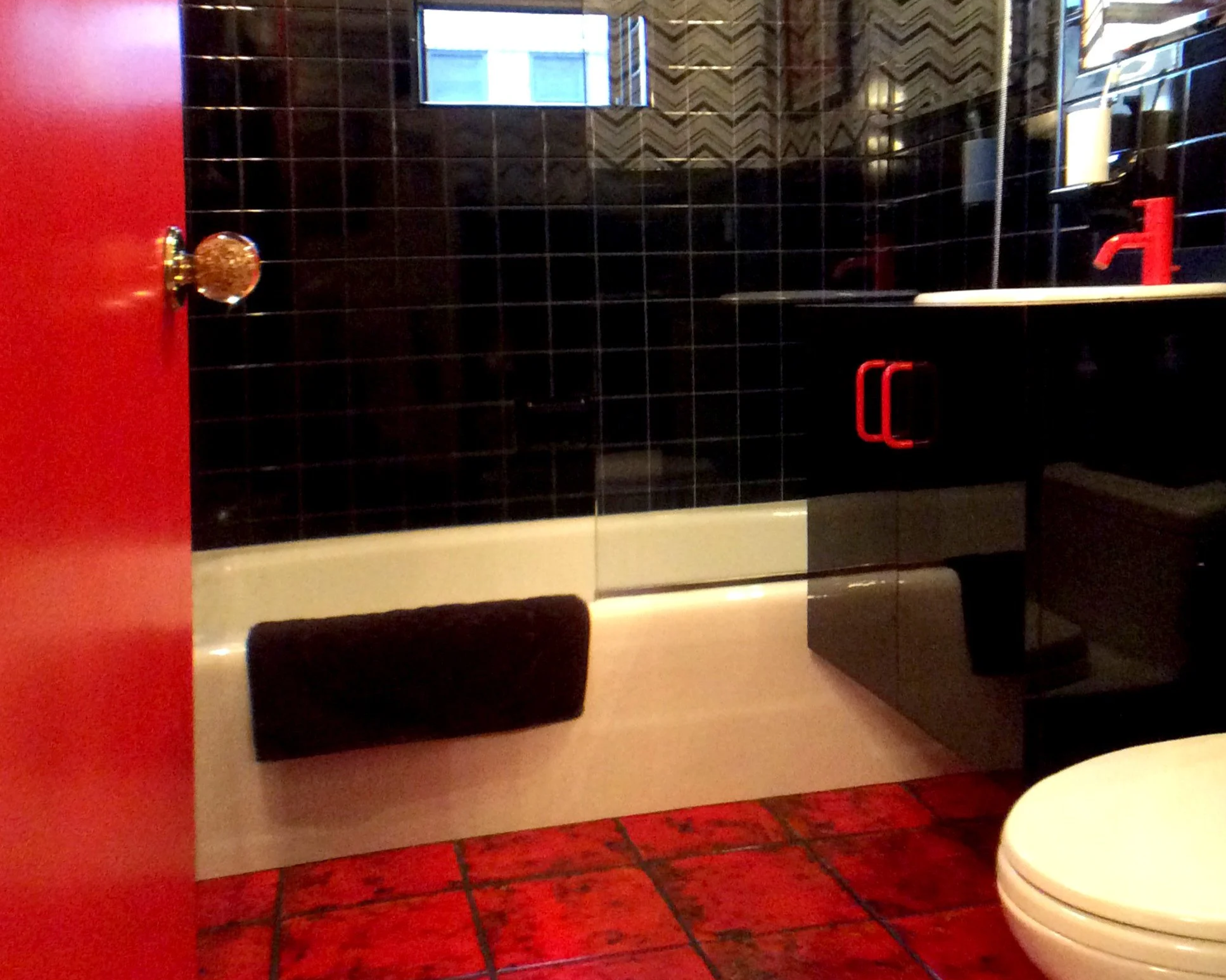 A mod 1970's bathroom with a black-tiled wall, a red door with a gold doorknob, and a black vanity with red handles. There is a mirror and a window with a view of a building outside. The floor is red-tiled, and a toilet is partially visible on the ri