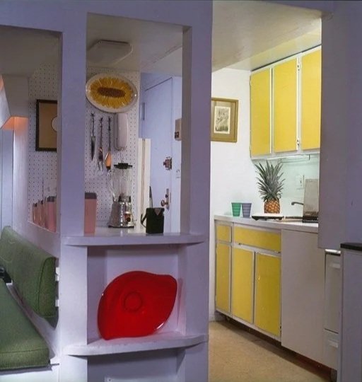 View of a kitchen with yellow and white cabinets, a pineapple on the countertop, and two decorative green cups. Part of a dining nook with a green banquette and a red glass dish on a shelf is visible in the foreground.
