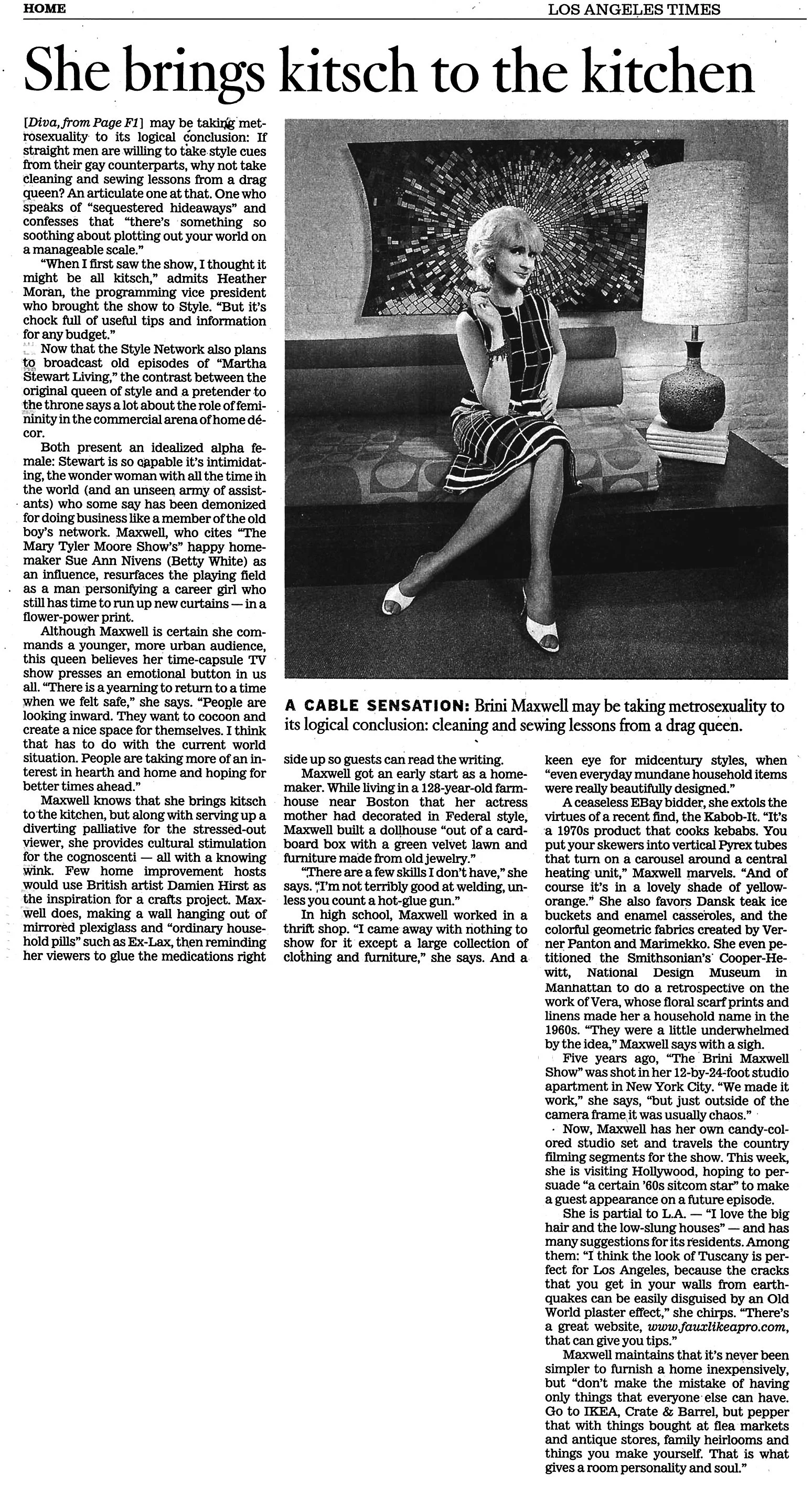 Black and white photograph of Brini Maxwell, sitting on a sofa in her living room. She is dressed in a plaid dress, wearing heels, and has short, styled hair. Behind her is a large, decorative wall piece and a table lamp.